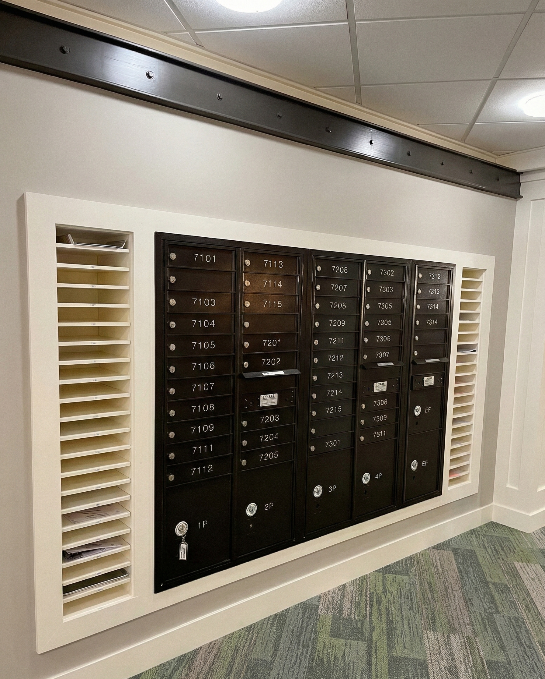 Wall-mounted black mailbox unit with labeled individual compartments, surrounded by white shelving and a carpeted floor.