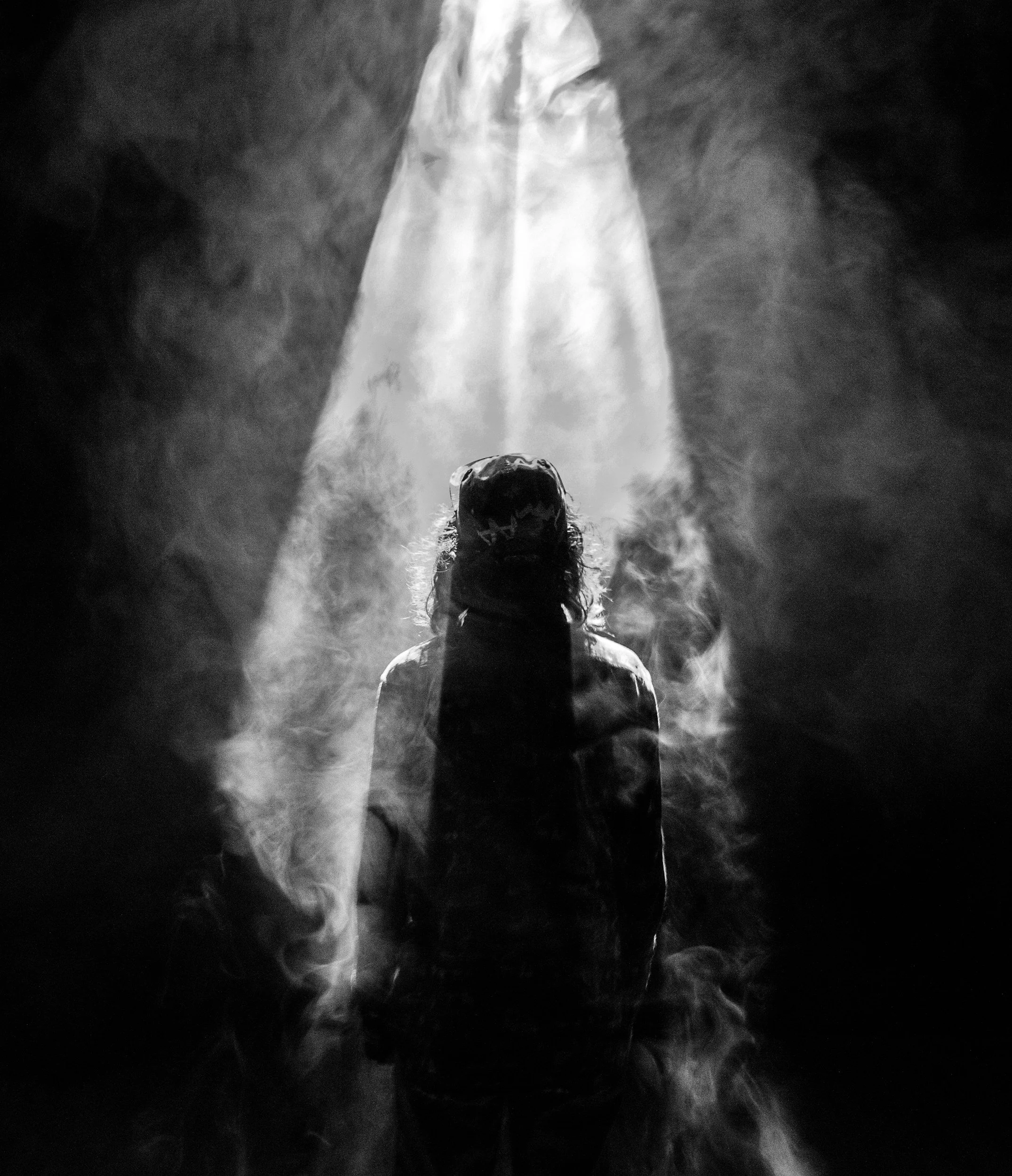 A person standing in a dark space with their back to the camera, illuminated by a bright beam of light coming from above, with smoke or fog surrounding them.