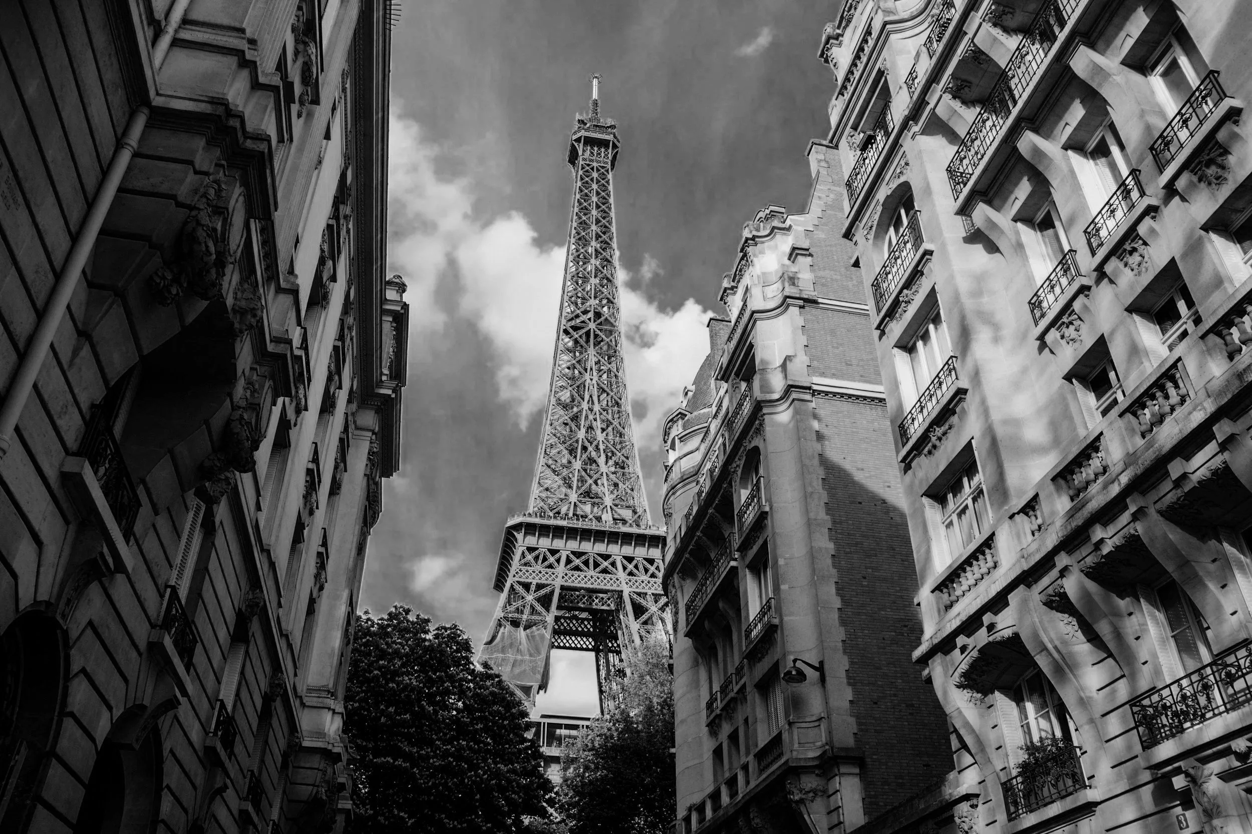 Black and white photo of the Eiffel Tower framed by tall Parisian buildings with balconies, viewed from the street level.