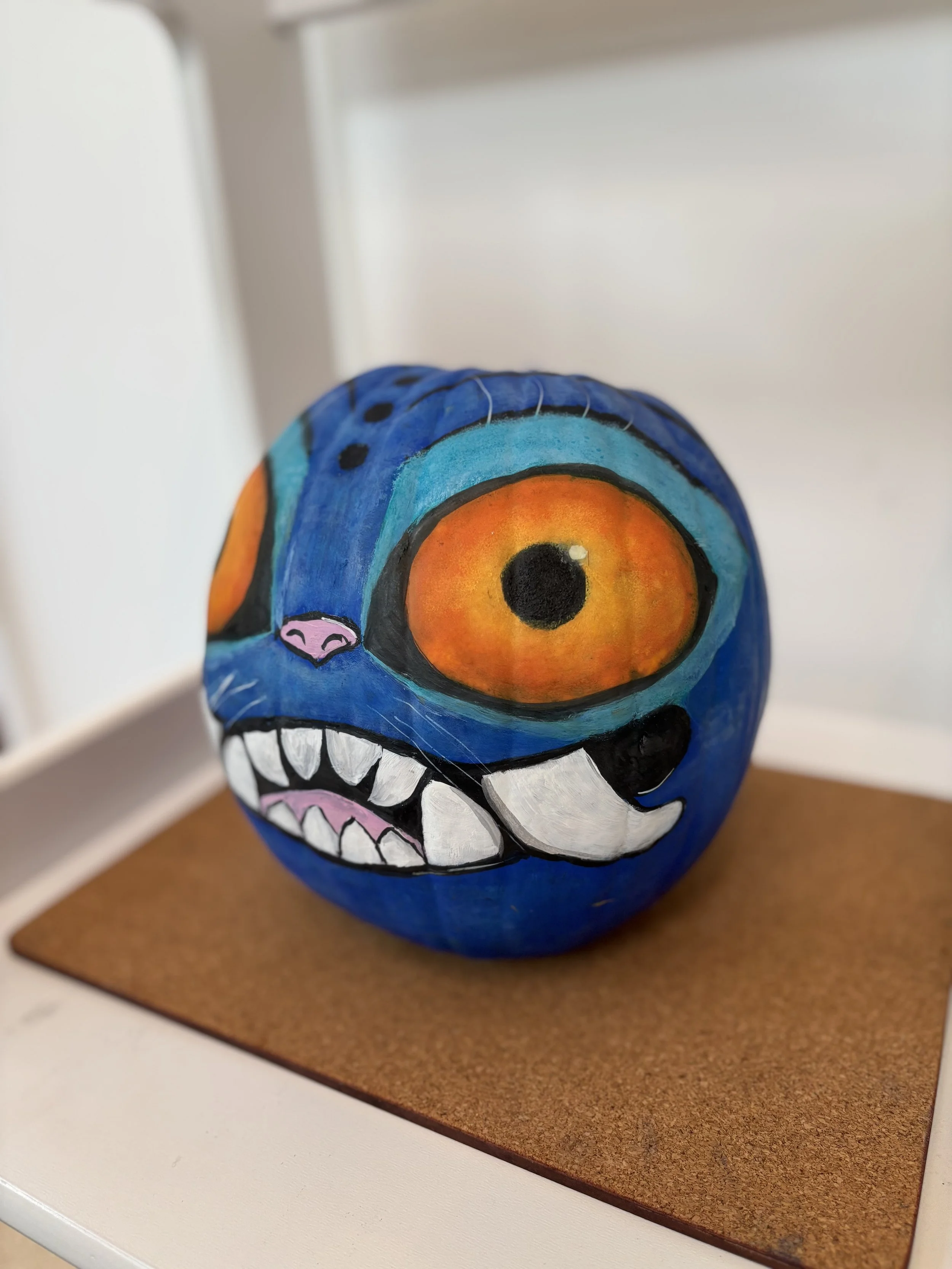 A painted rock with a colorful, monster face design, featuring large orange eyes, sharp teeth, and a pink nose, placed on a cork mat.