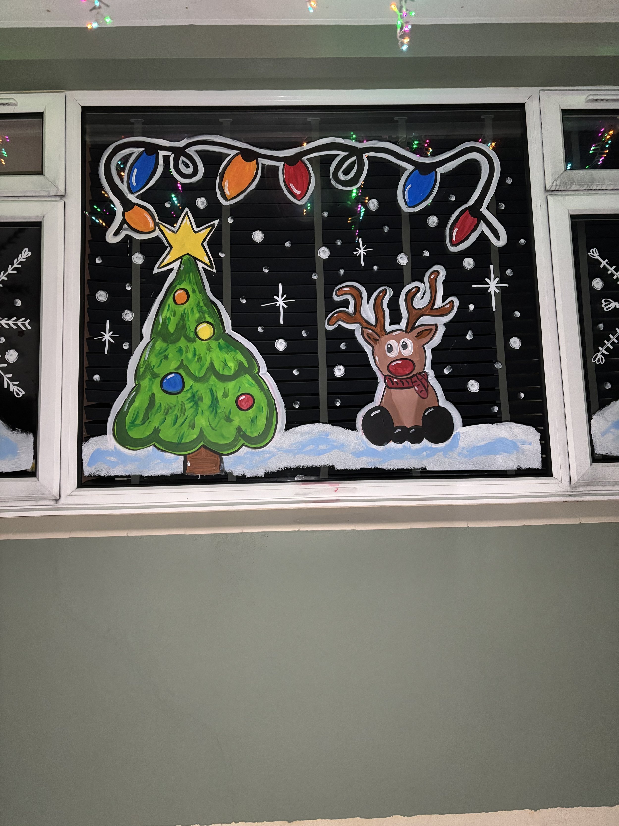 Decorative Christmas window with painted decorations of a Christmas tree with multicolored ornaments and a yellow star on top, a reindeer with big eyes and red nose, snow on the ground, white snowflakes, and Christmas lights hanging overhead.