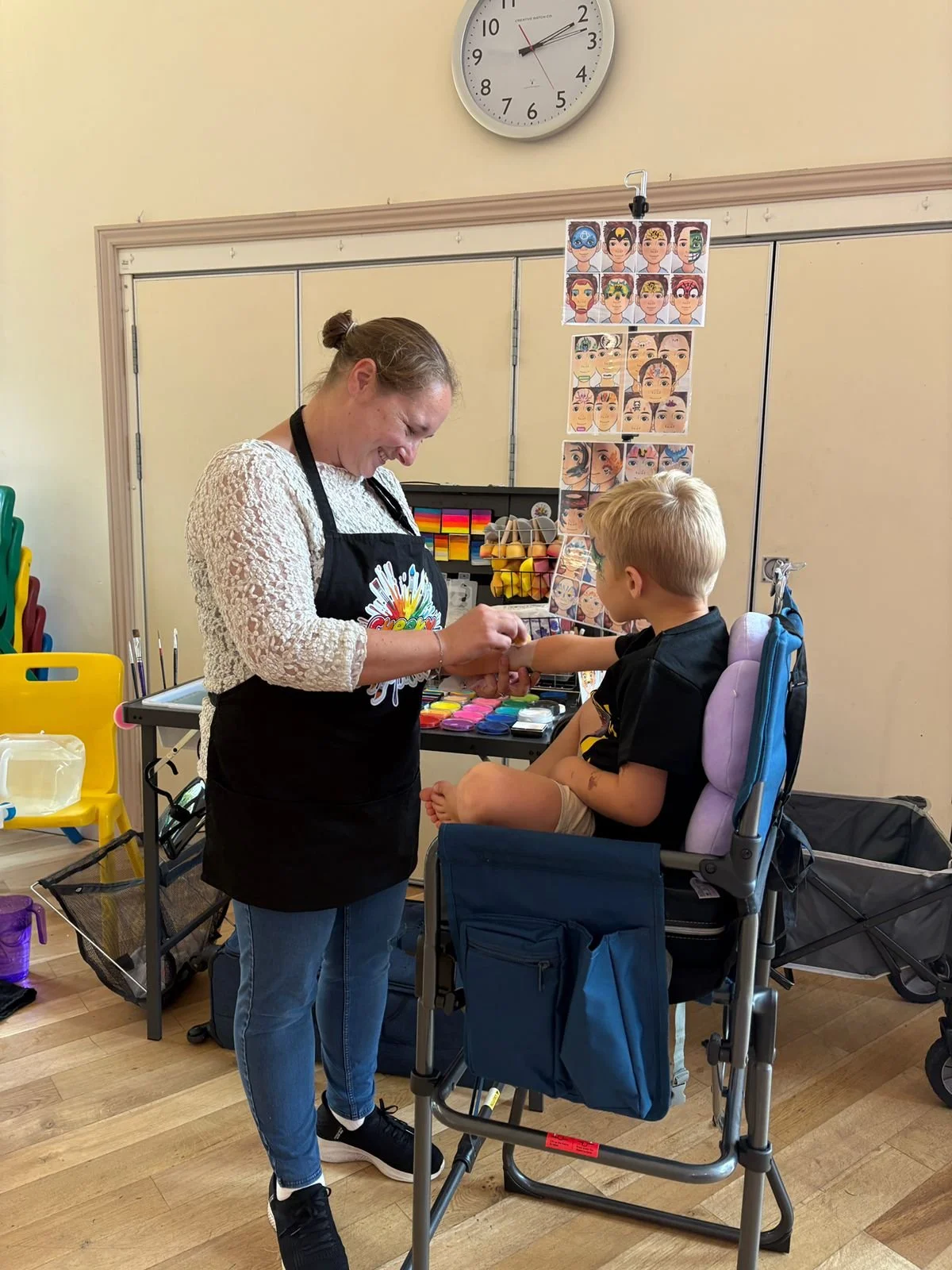 A woman giving a manicure to a young boy sitting in a specialized chair, with colorful face painting and makeup supplies on nearby tables, in a room with yellow walls and a clock showing 10:11.