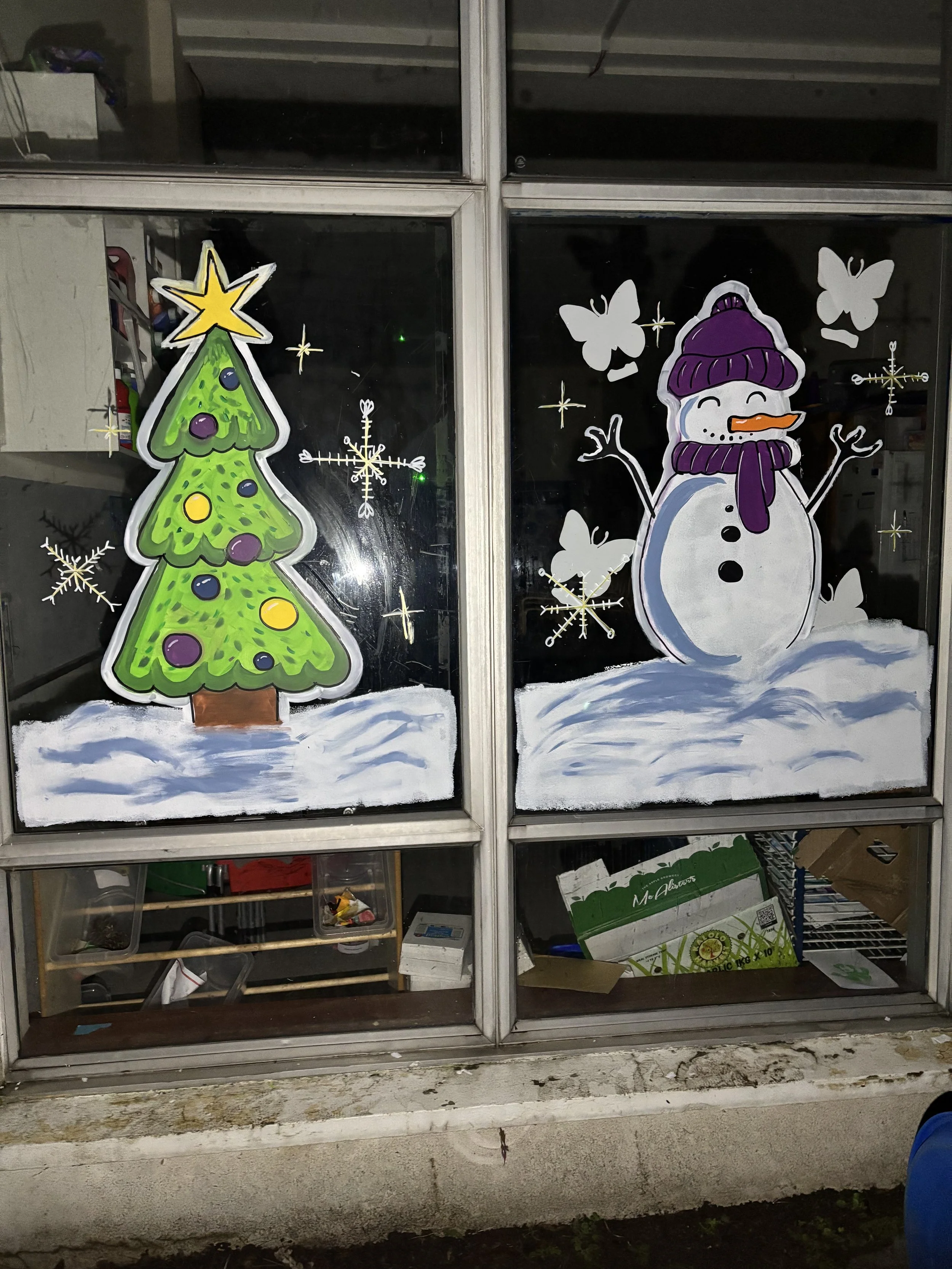 Decorative Christmas window art featuring a Christmas tree with colorful ornaments and a star on top on the left, and a snowman with a purple hat, scarf, and carrot nose on the right, with snowflakes and butterflies around them.