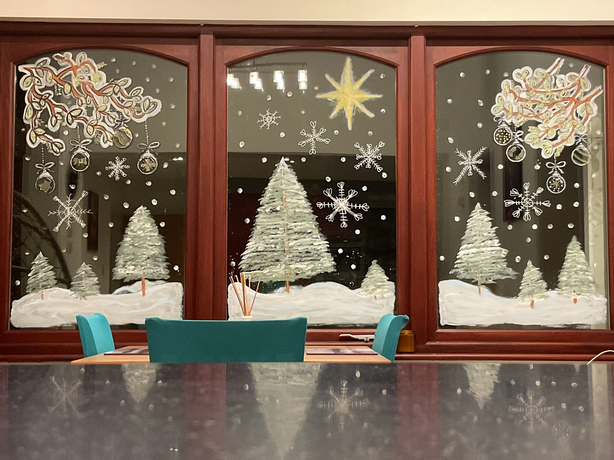 Decorated window with white Christmas trees, snowflakes, ornaments, and stars, viewed from inside a room, with a table and teal chairs in the foreground.