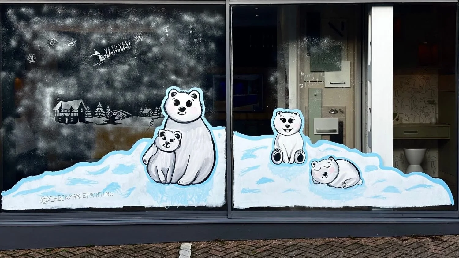 Window mural painted with adorable polar bears on snow with a wintery landscape background, including houses and trees.