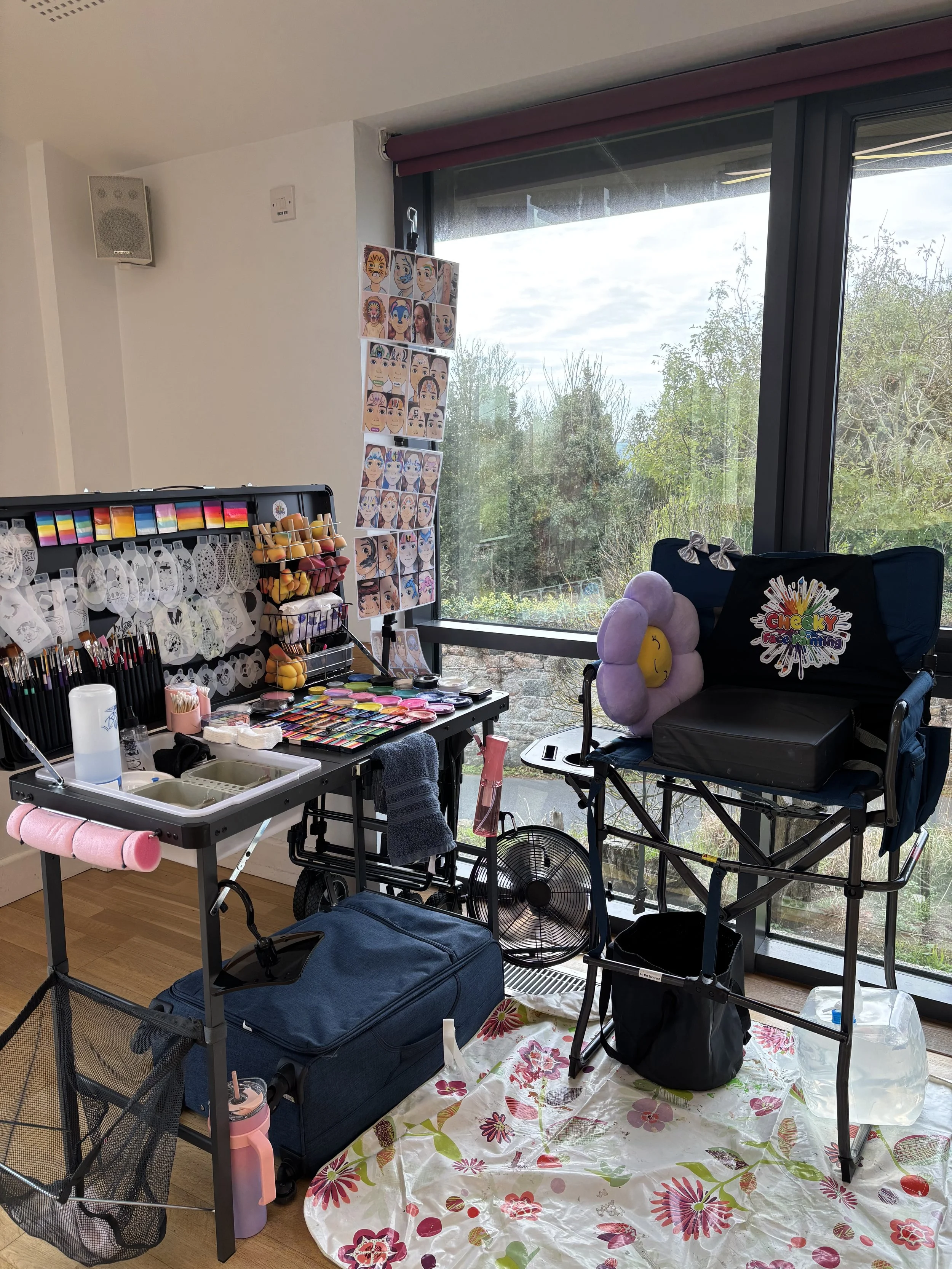 Indoor face painting station with colorful supplies, makeup palettes, brushes, and painted face designs on hanging display, with a large window showing trees outside.