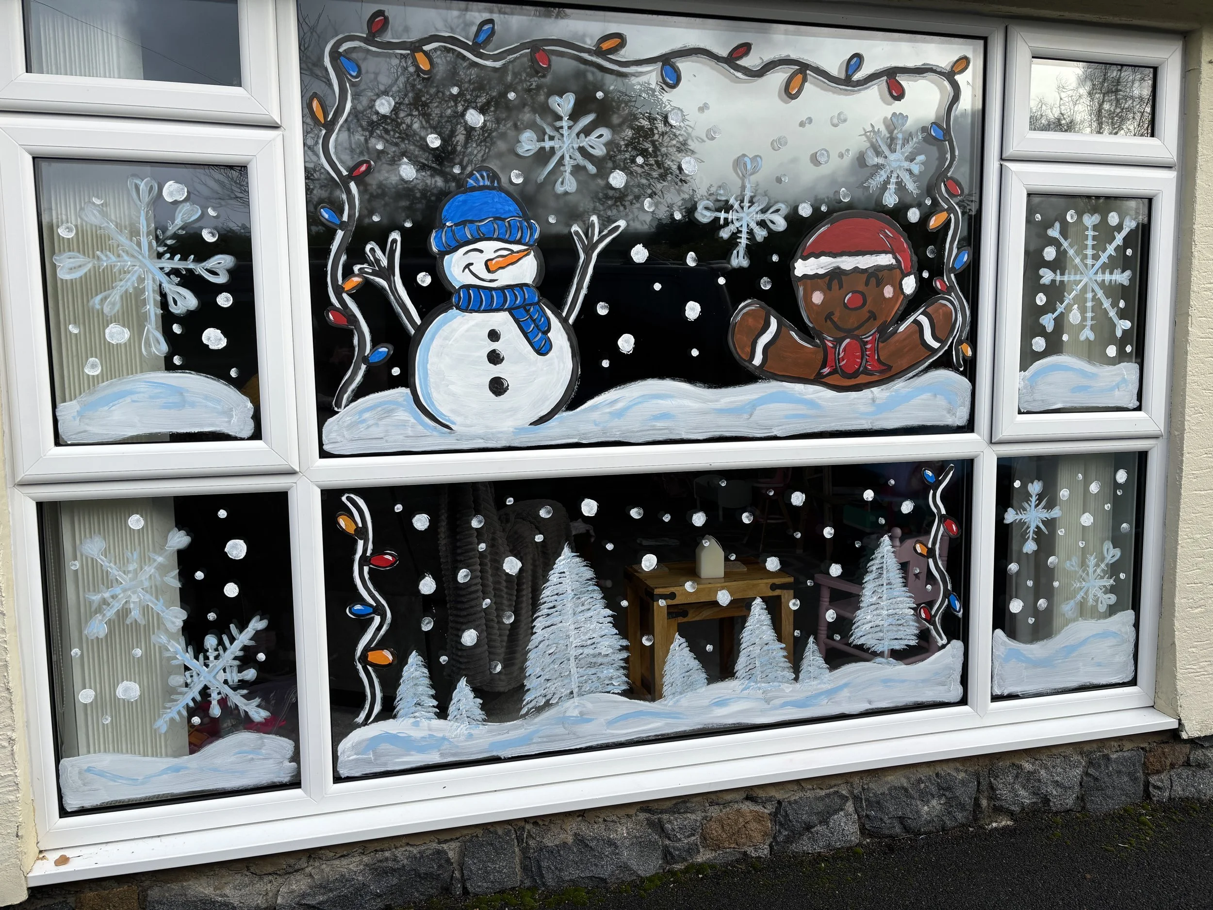 Decorated window with snowman, gingerbread man, snowflakes, Christmas lights, pine trees, and snow.
