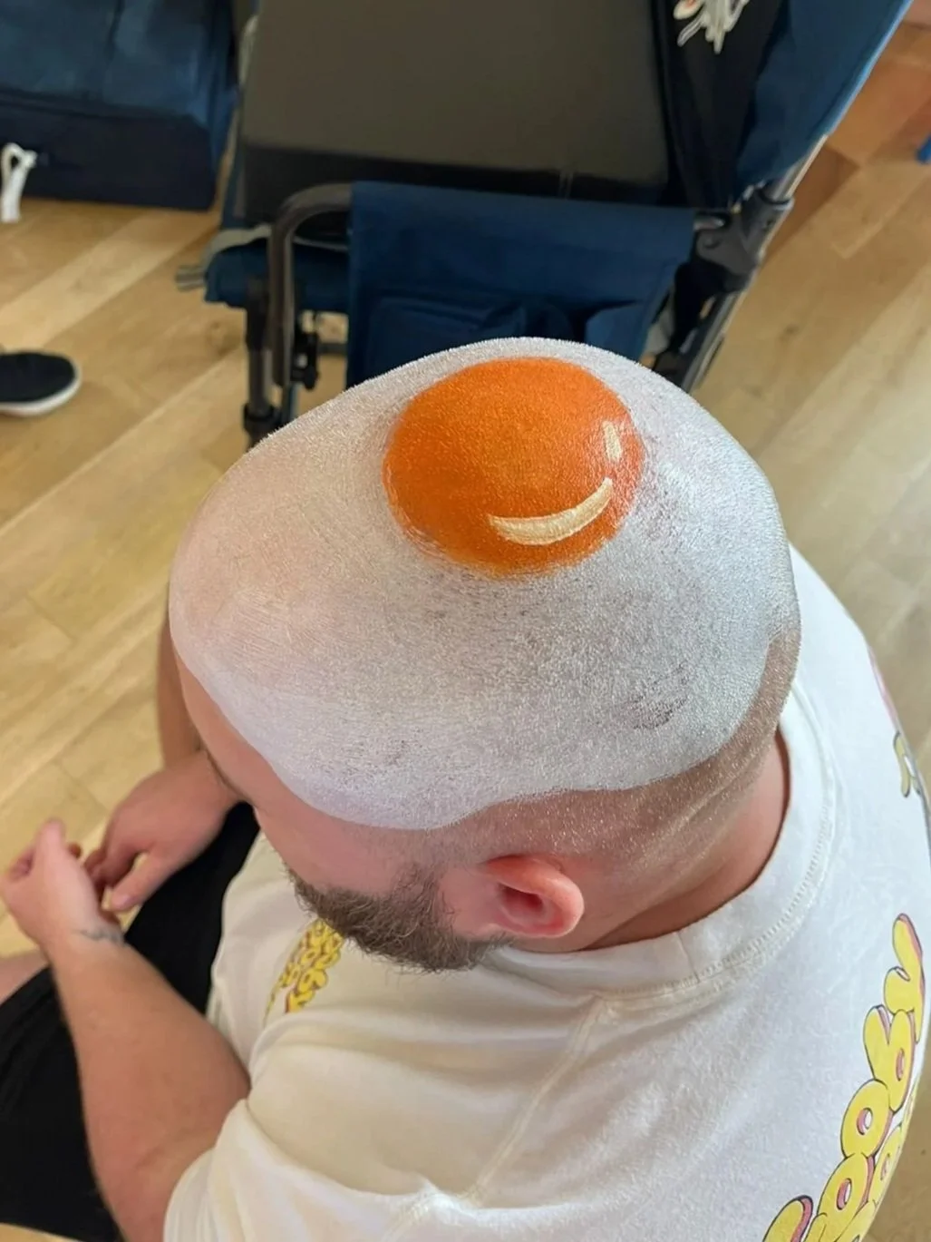 Top view of a person's shaved head with a smiley face drawn using orange hair dye, showing a smile and eye, and a stencil mask for hair coloring.