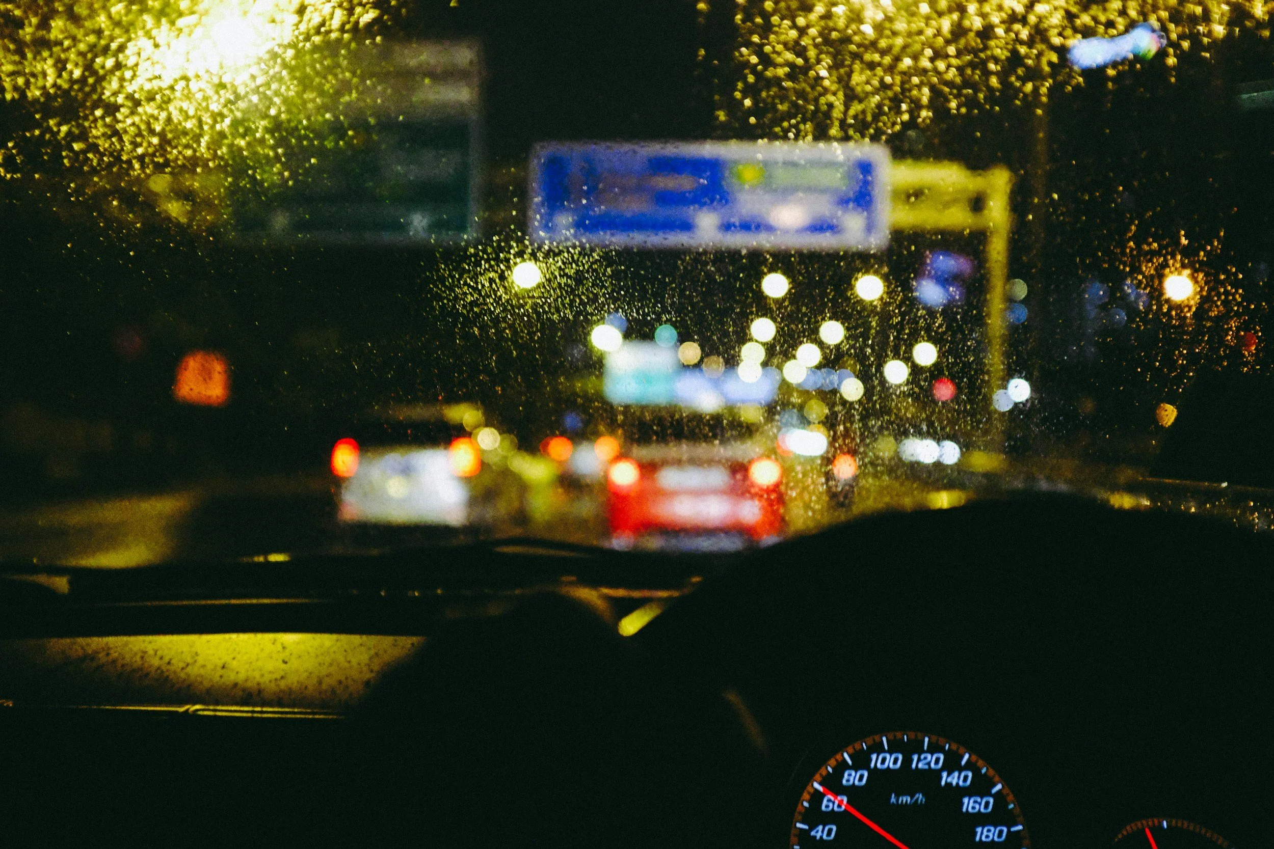 View from inside a car looking through a rain-covered windshield at wet city streetlights and traffic at night, with illuminated signs and vehicles ahead.