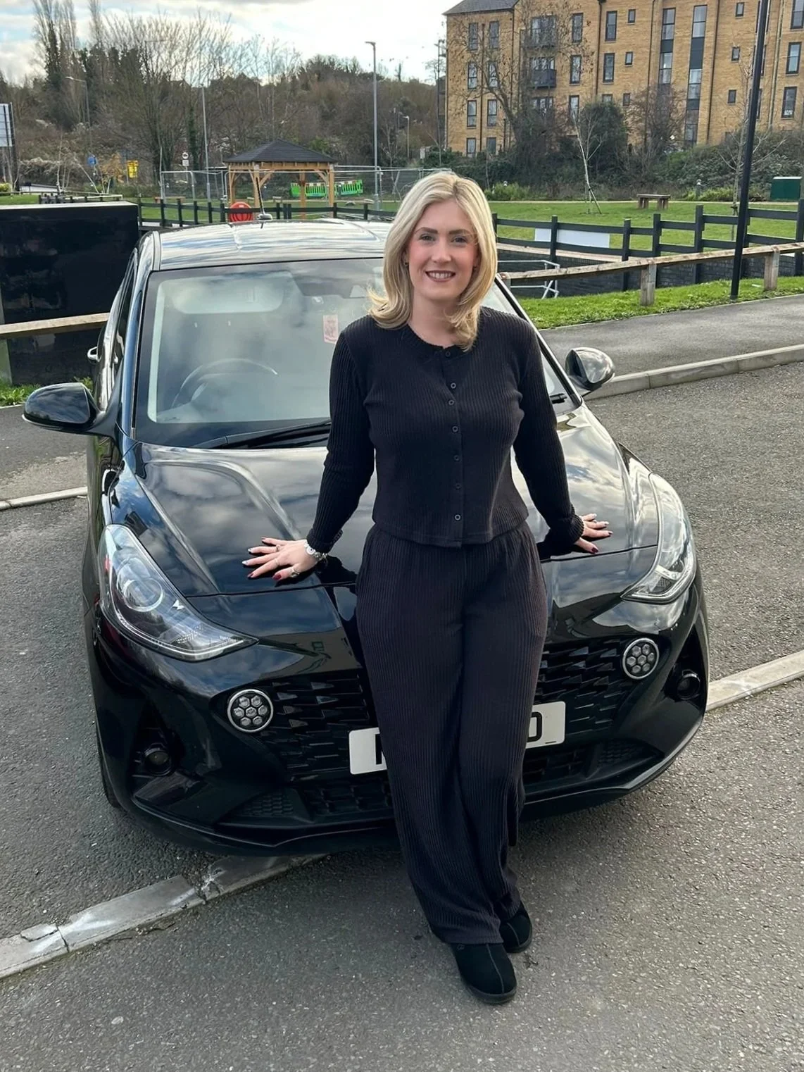A woman with blonde hair, wearing a black ribbed sweater and matching black pants, stands in front of a black car in a parking lot outdoors. She is smiling and posing with her hands resting on the car's hood.