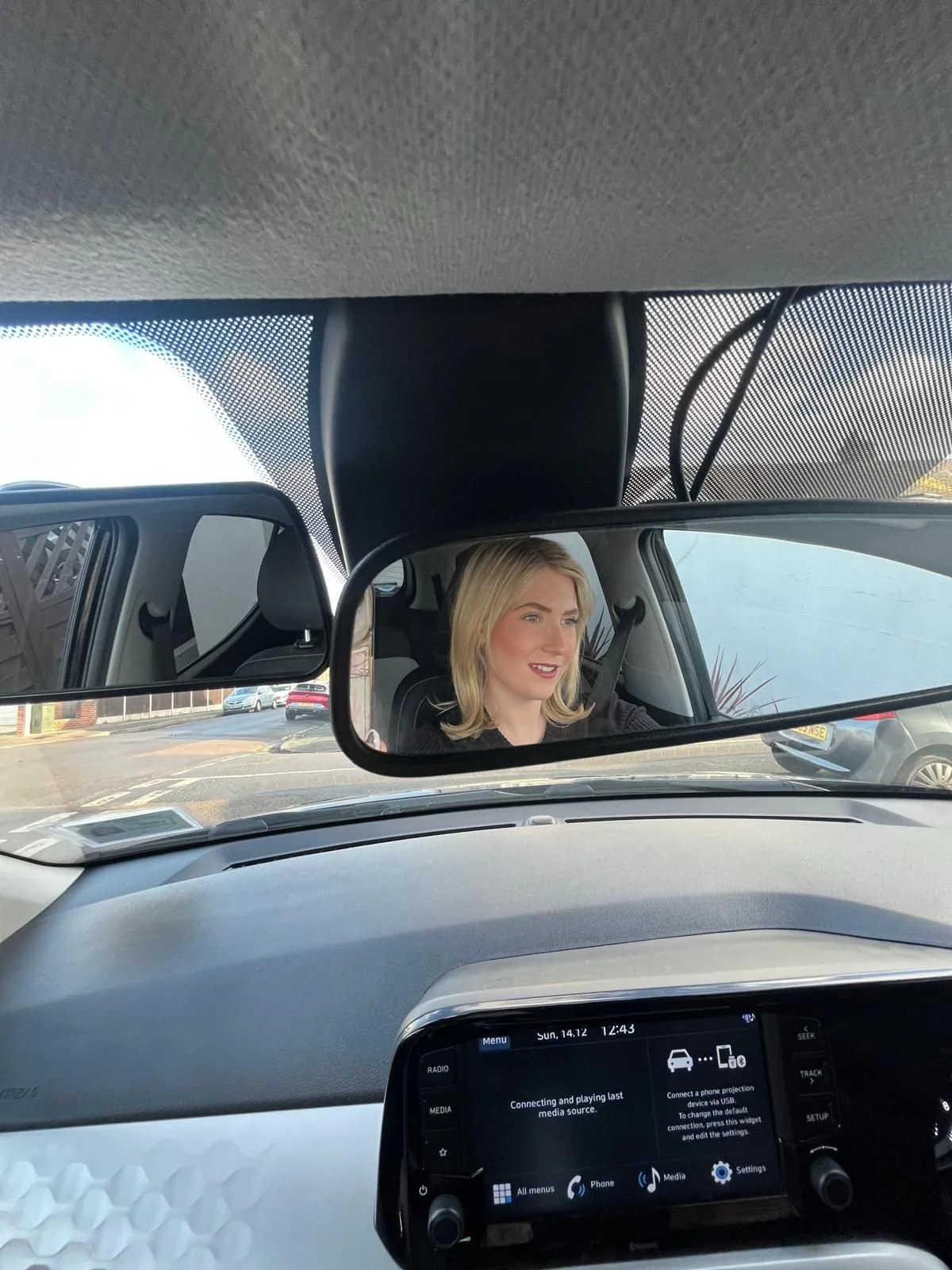 A woman with blonde hair sitting in a car, looking at the rearview mirror. The car's dashboard and part of the interior are visible, with a parking lot and buildings outside the car window.