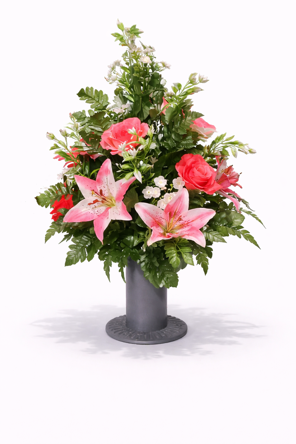 A floral arrangement in a dark gray vase featuring pink lilies, pink roses, white small flowers, and green foliage on a white background.