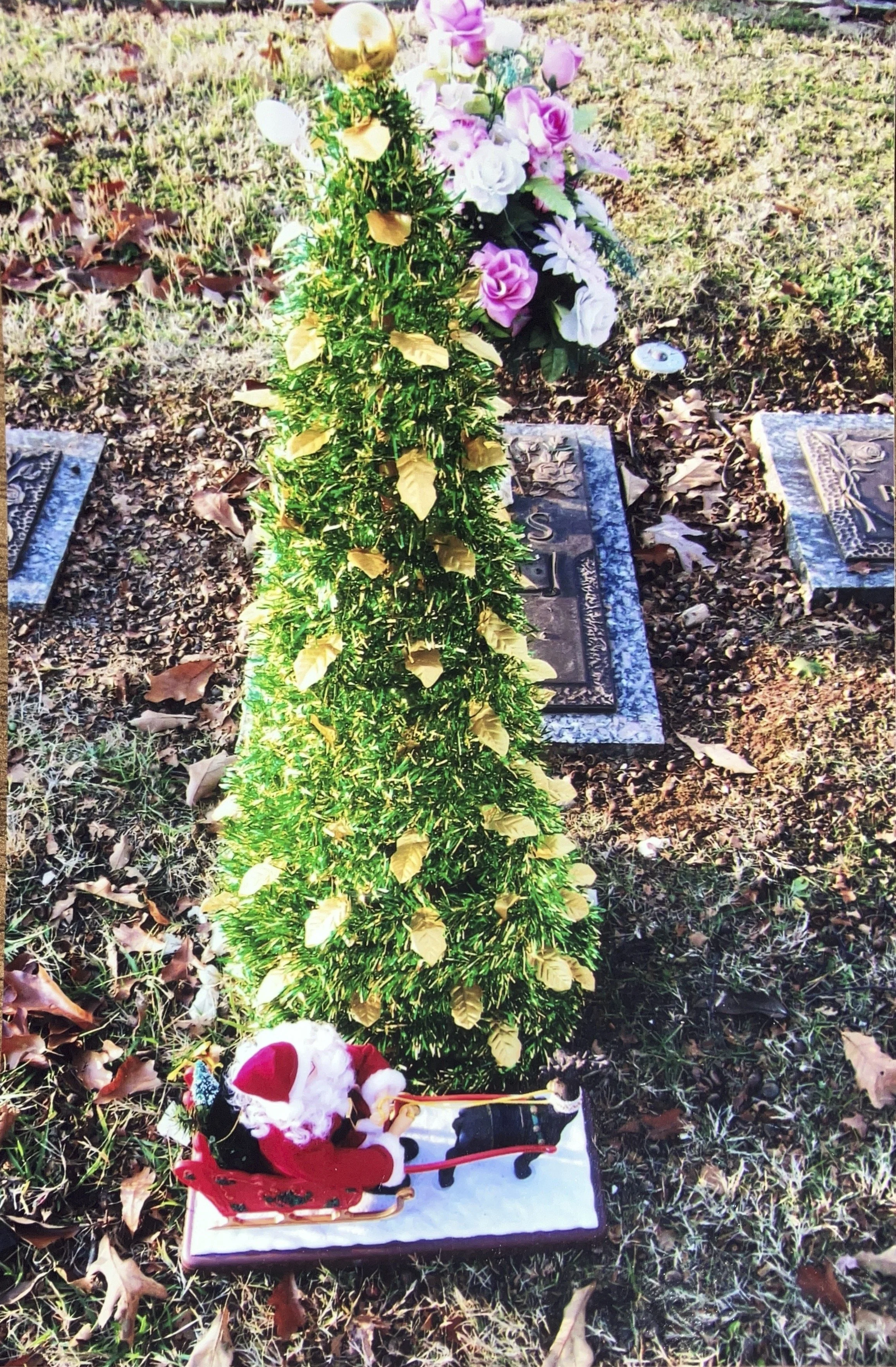 Small green Christmas tree with gold leaves on a cemetery grave site, with a Santa Claus figurine riding a sleigh at the base, and a bouquet of pink and white flowers nearby, surrounded by fallen leaves.