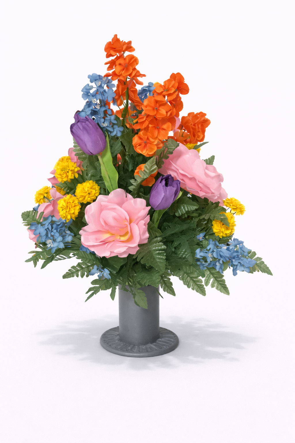 Artificial floral arrangement in a gray vase, featuring pink roses, purple tulips, orange flowers, yellow blooms, and blue flowers, with green foliage.