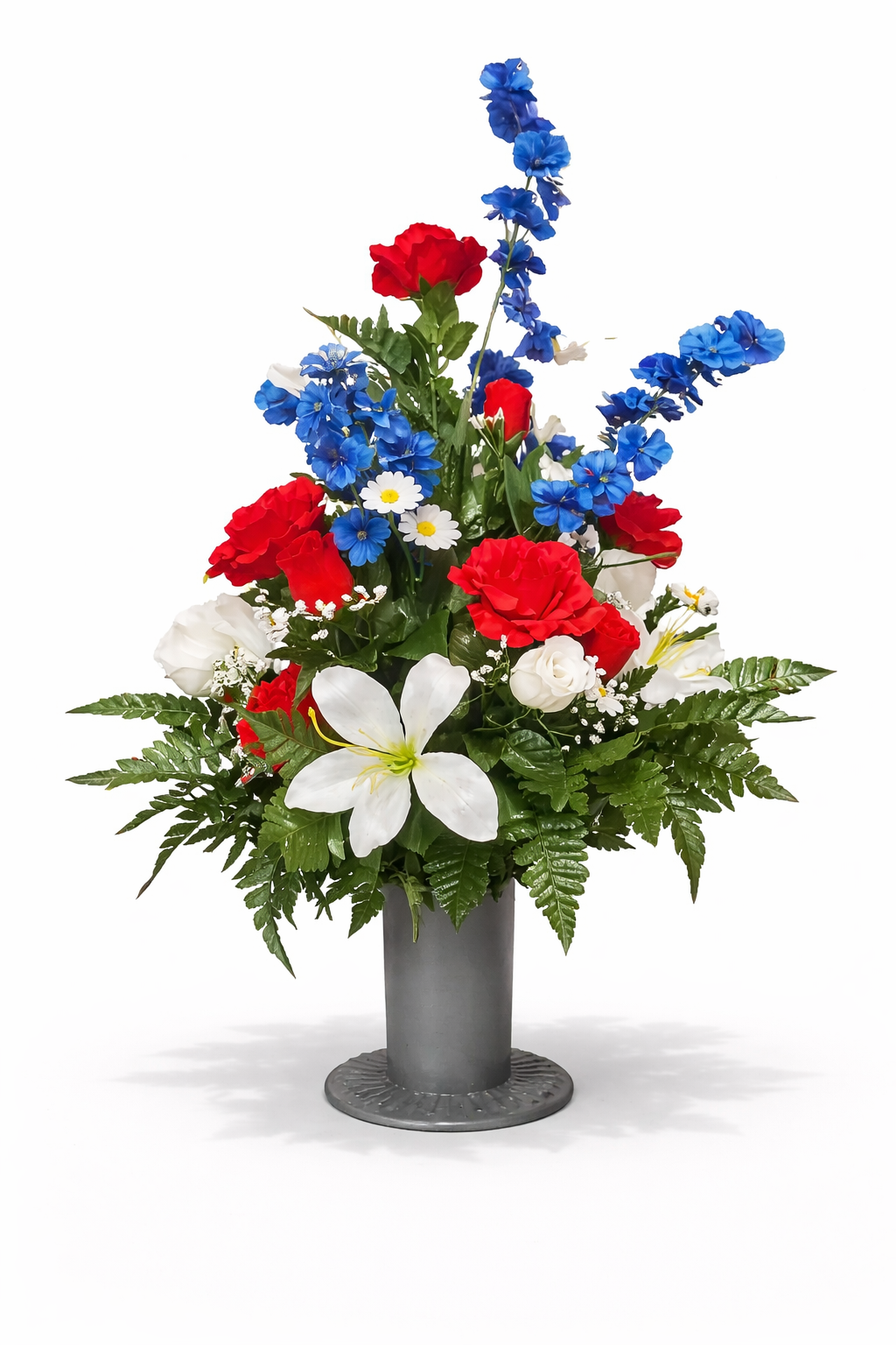 A bouquet of red, white, and blue flowers in a gray vase, including lilies, daisies, and delphiniums, arranged with green foliage.