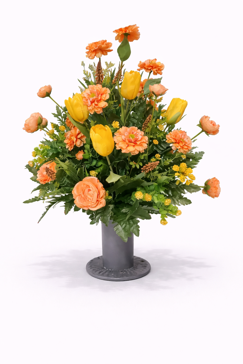A colorful flower arrangement in a gray vase with yellow tulips, orange carnations, and various green foliage on a white background.