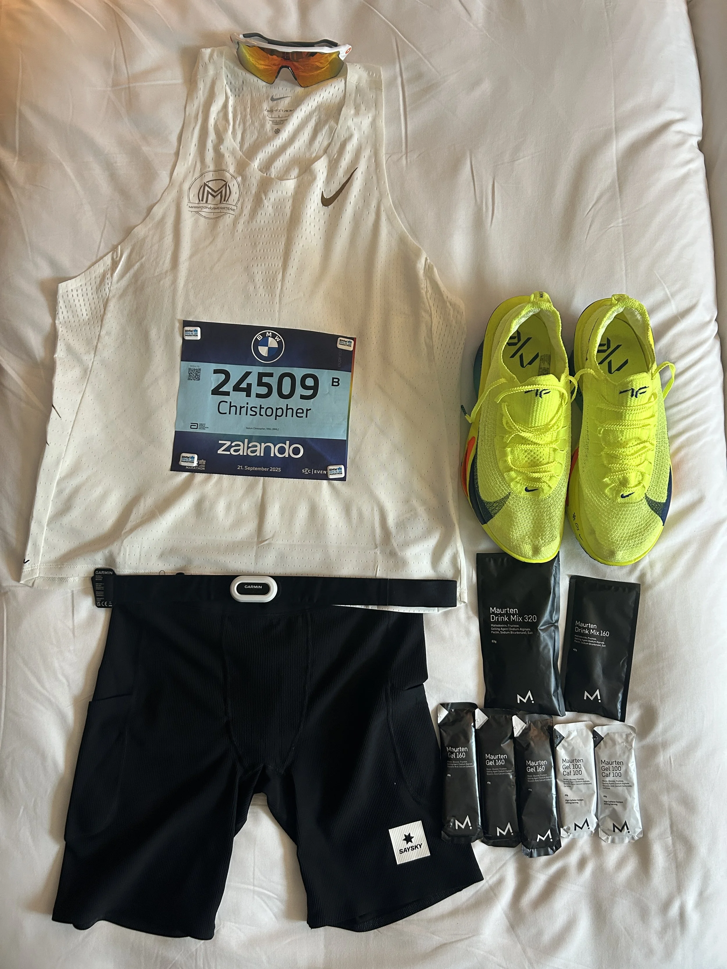 En samling av løpetøy og tilbehør, inkludert en hvit løpe-vest med Nike og Maratonmerket, et startnummer med navnet Christopher, løpesko i neon gul, en svart bukse, en pulsmåler, og pakker med Maurten gel for energi.