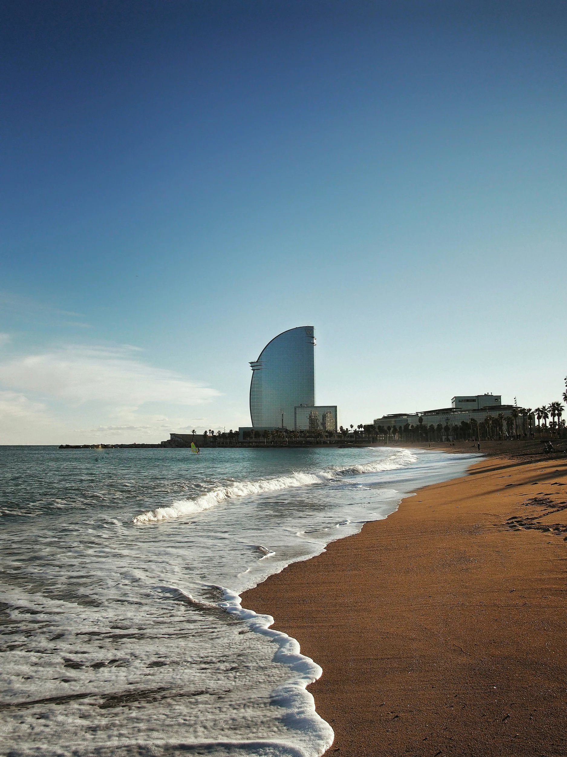Bilde av en strand med sand og hav, hvor bølger slår inn, og i bakgrunnen en moderne hotellbygning, som ser ut til å være W Barcelona.