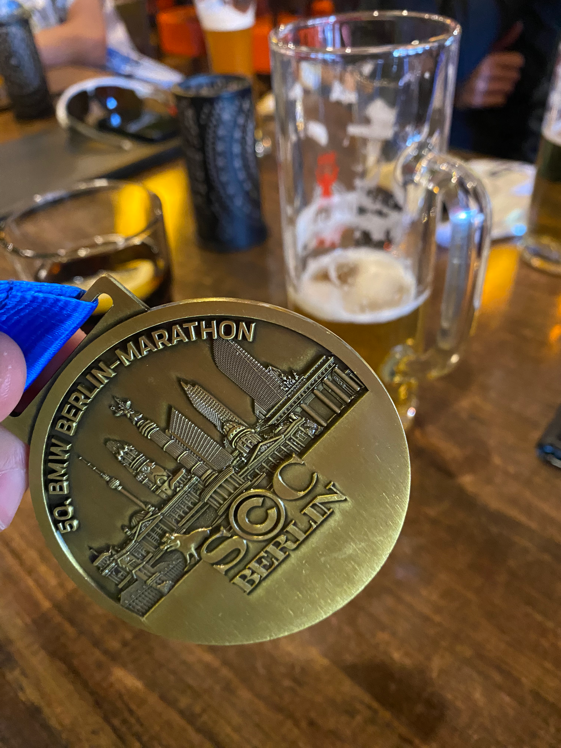 En person holder en gullmedalje med teksten '50. Berlin-Marathon' foran en halvt full ølbong. På bordet er det flere ølglass, en telefon, og noen mørke containere.