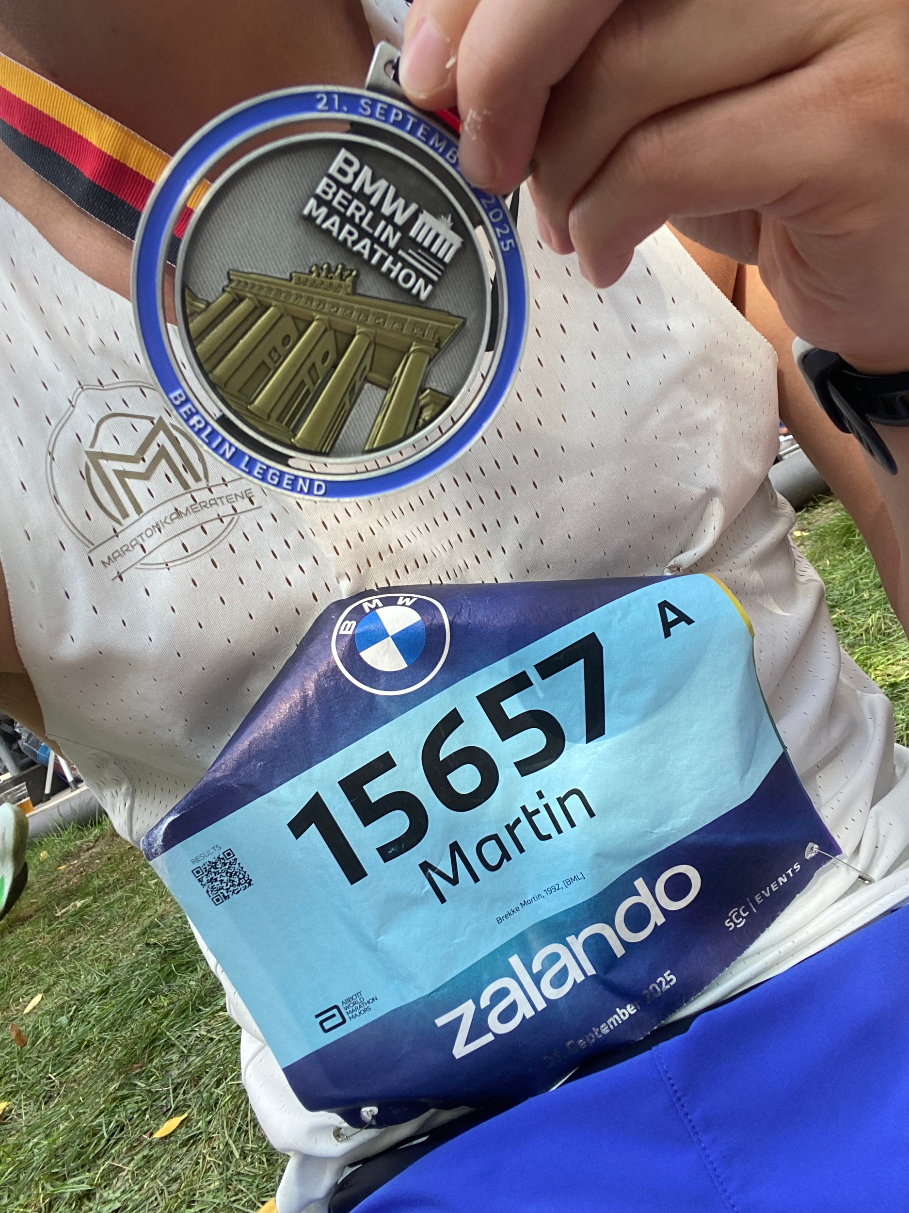 En person holder opp en finisher-medalje fra Berlin Marathon 2023, med en Berlin-legende på jakken og en startnummer på kroppen.