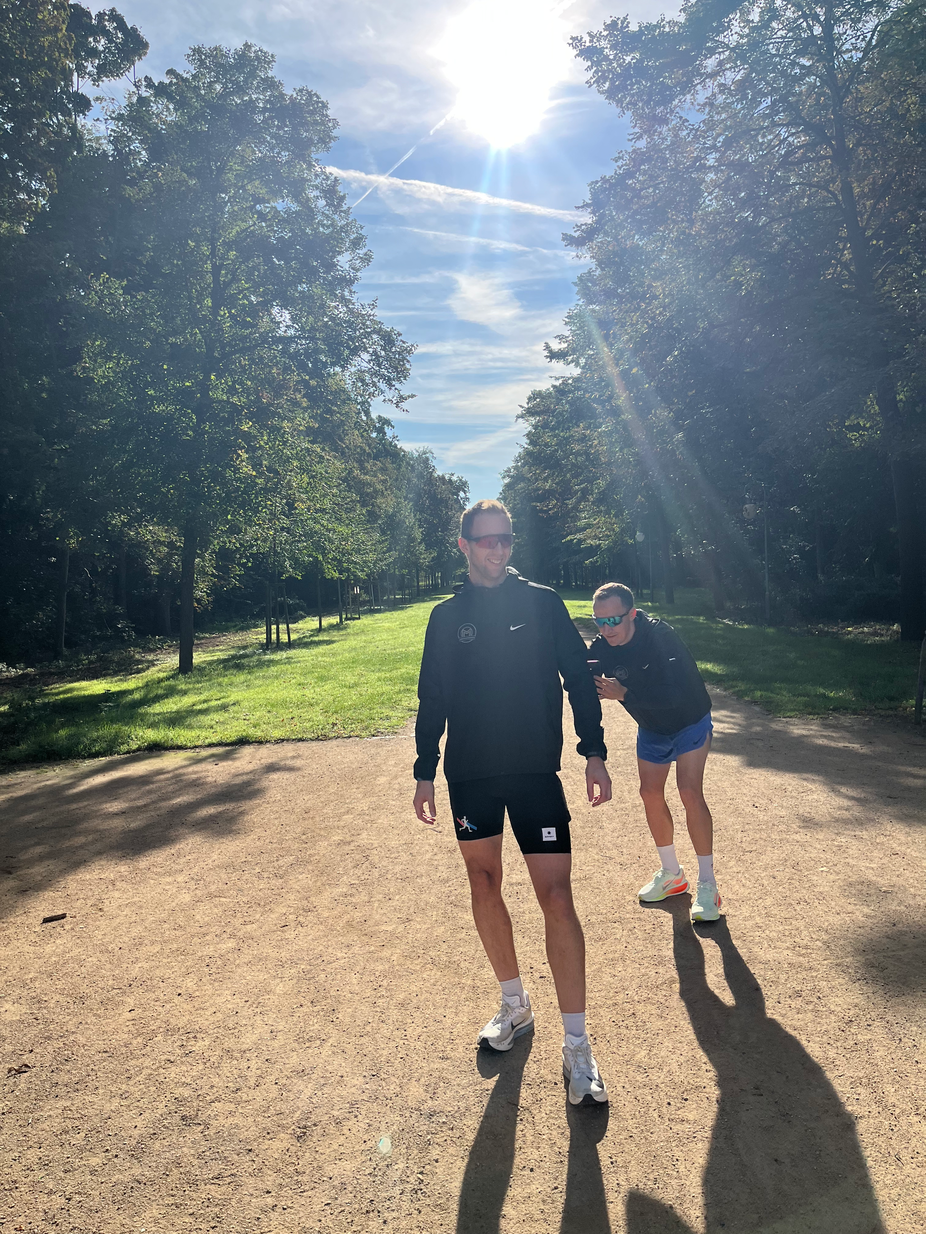 To mennesker går i en park under sollys, den ene personen smiler og kler seg i treningstøy, mens den andre ser på telefonen, alle med solbriller.