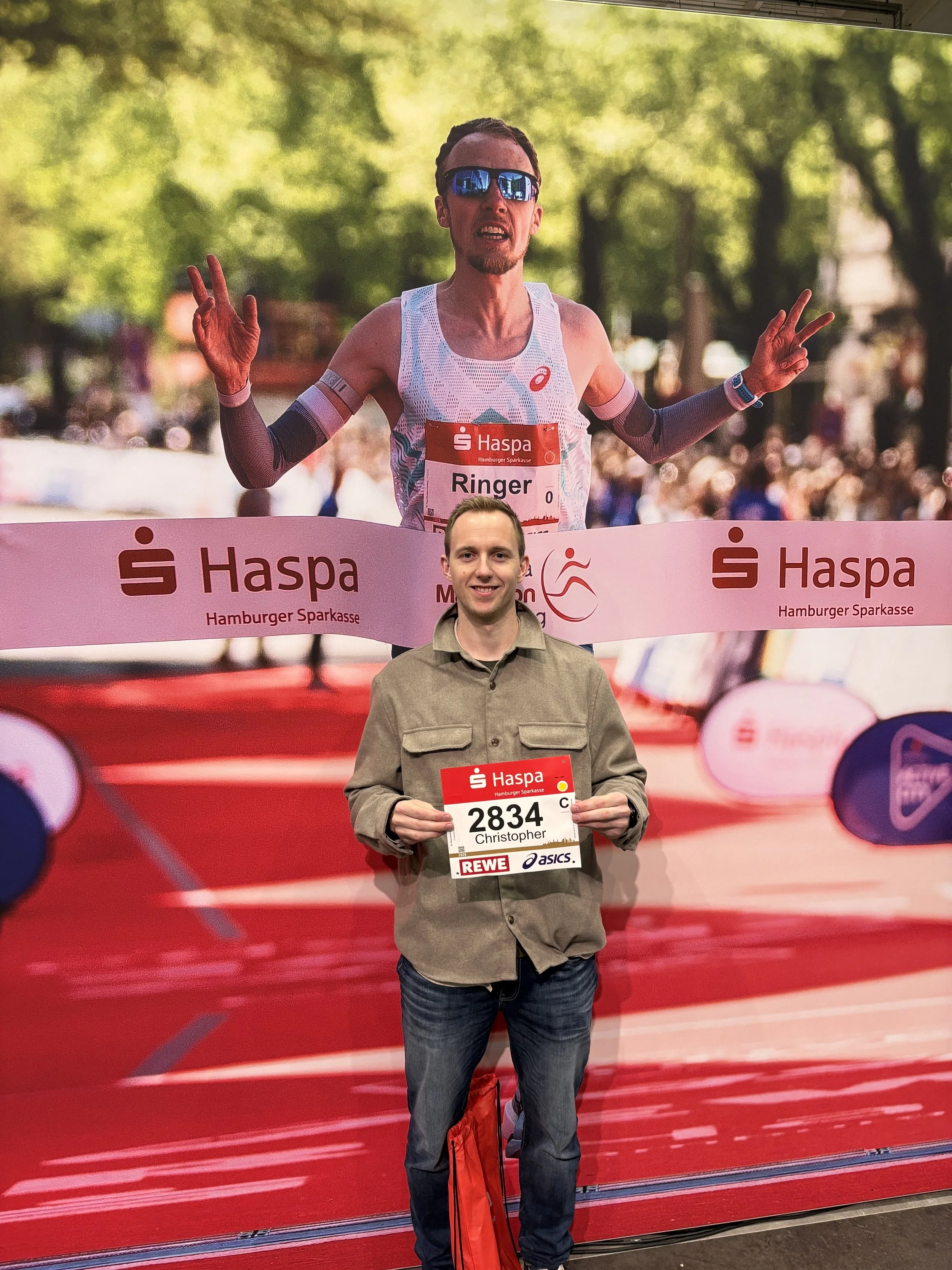 Person med startnummer 2834, som heter Christopher, poserer foran et bakgrunnsbilde av en maratonløper under et løp. Personen holder et startnummer og smiler.
