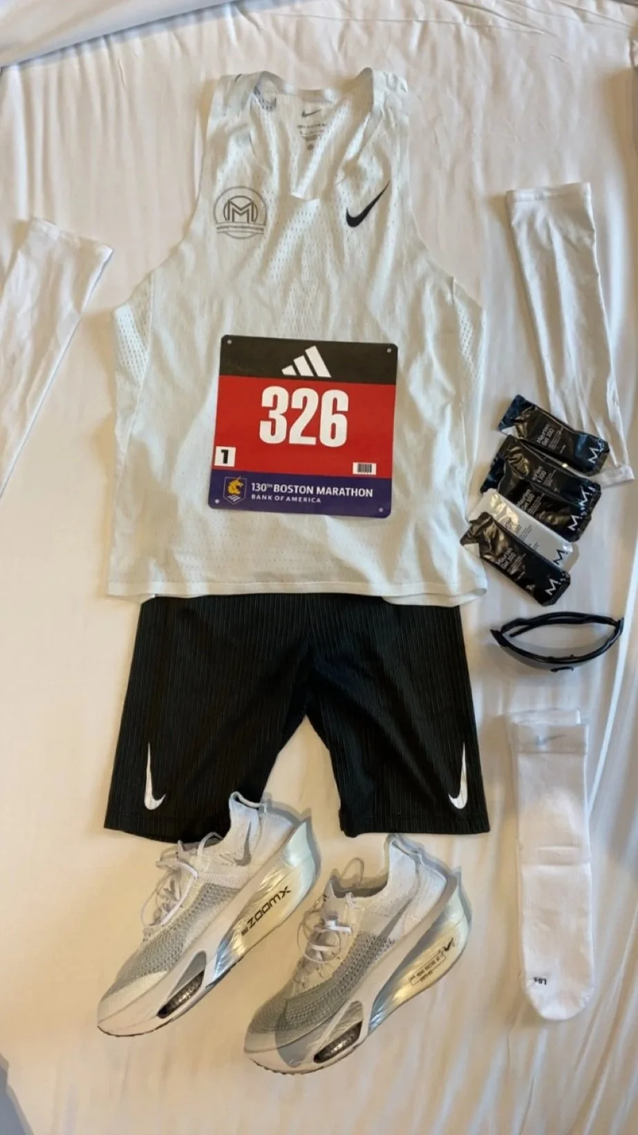 Løpeutstyr for marathon, inkludert løpebib, t-skjorte, løpebukser, løpesko, energibarer, solbriller og armbånd, lagt ut på seng.