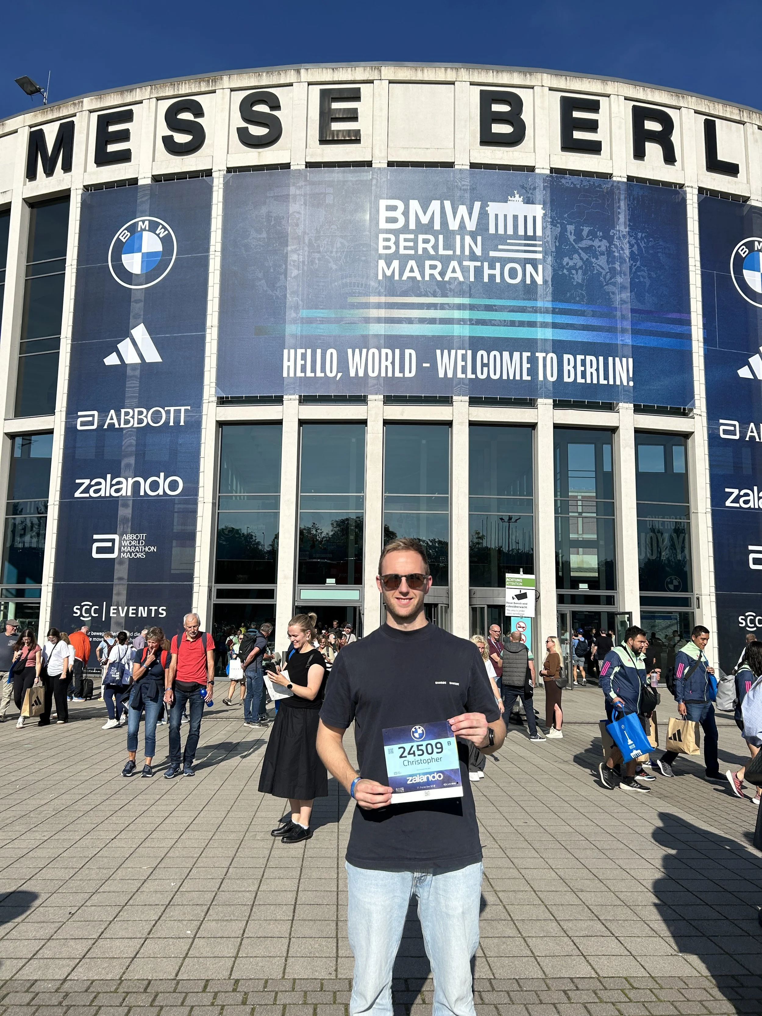 En mann som holder et deltakerbevis foran Messe Berlin, med mange mennesker i bakgrunnen, og en stor banner som reklamere for BMW Berlinmarathon.
