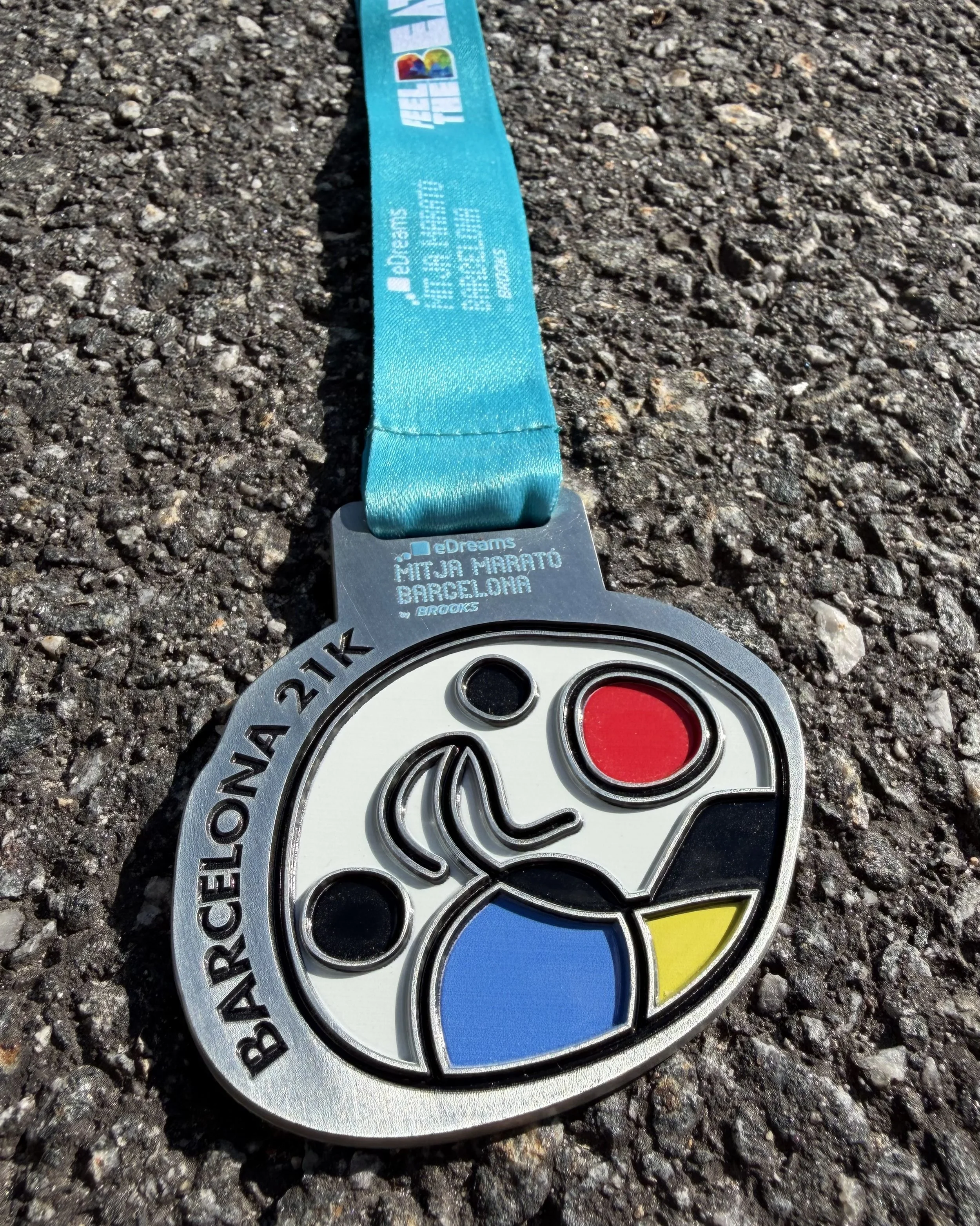 Et metallmedalje med fargerik rund logo og blått bånd, plassert på asfalt. Tekst på medallen inkluderer URL og navn på en løpsløype i Barcelona, samt sponsorer.