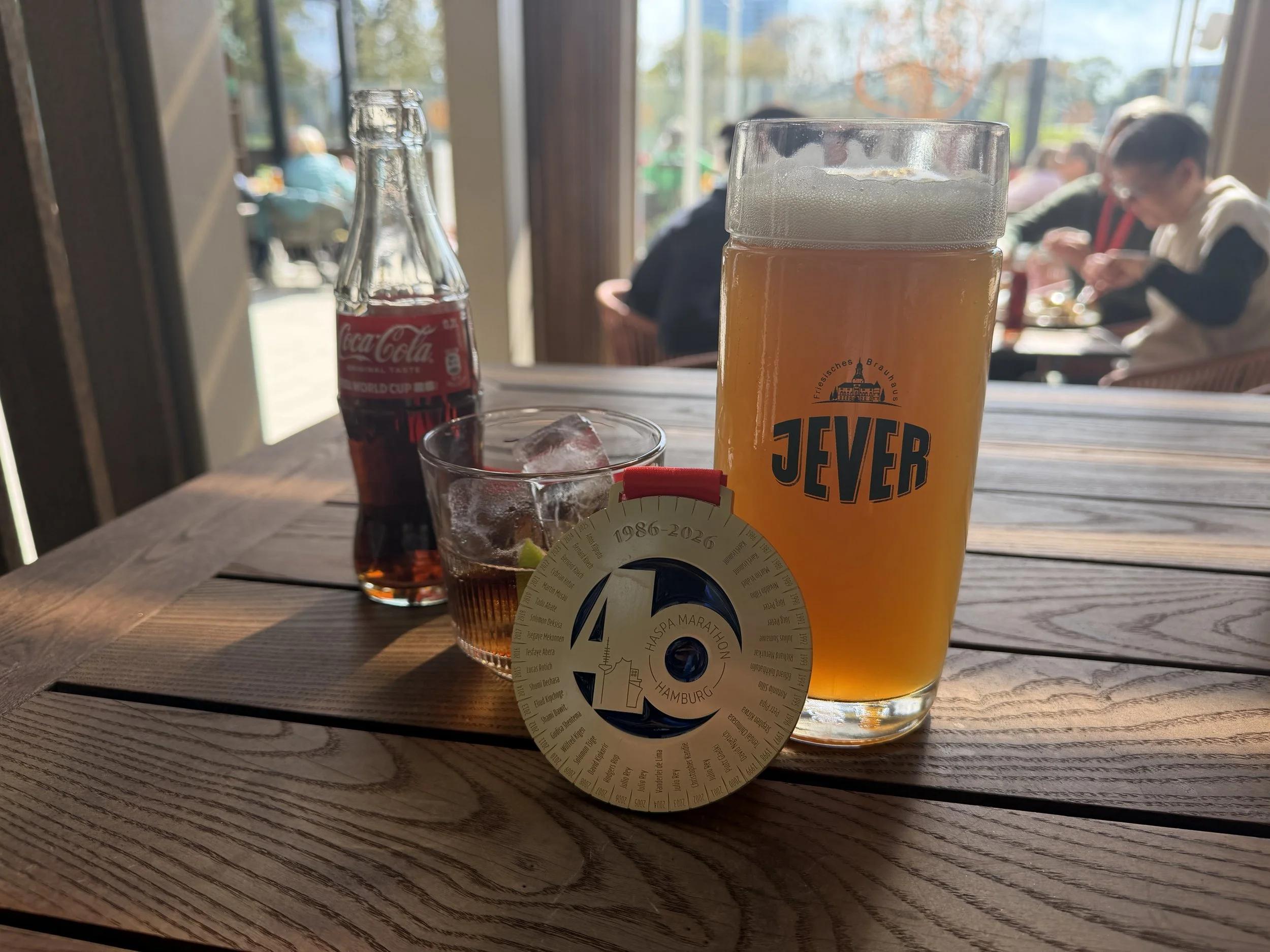Et glass øl med skum, en liten spritflaske Coca-Cola, en liten glass med is og lime, og et papirmerke med teksten »Hamburg 40» på en trebord i en restaurant eller kafé, med mennesker i bakgrunnen ved vinduer.
