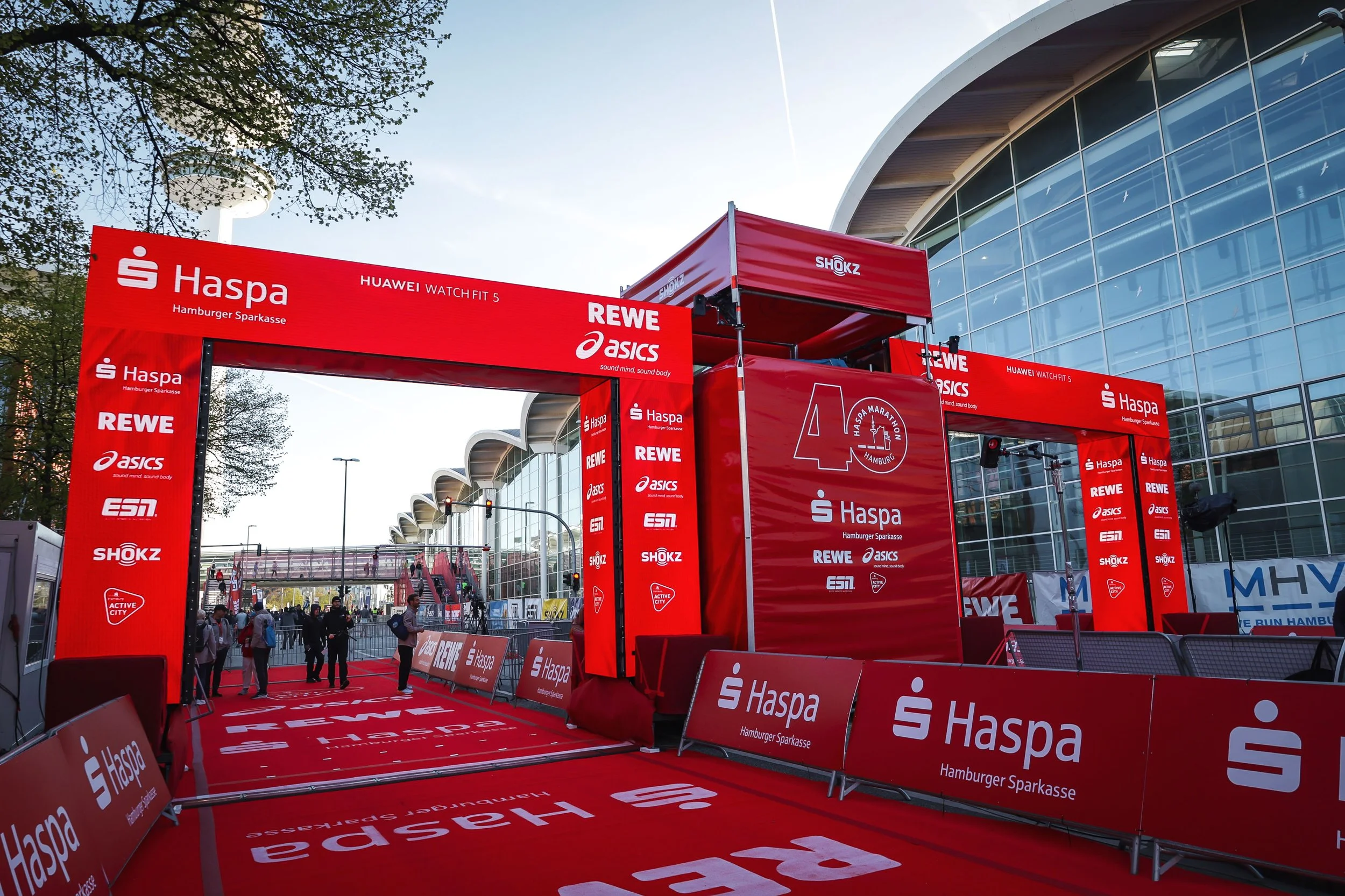 Innendørs løpsområde med stort rødt oppsett for Haspa Hamburg Marathon, omgitt av søte mennesker og med moderne glassfasader.