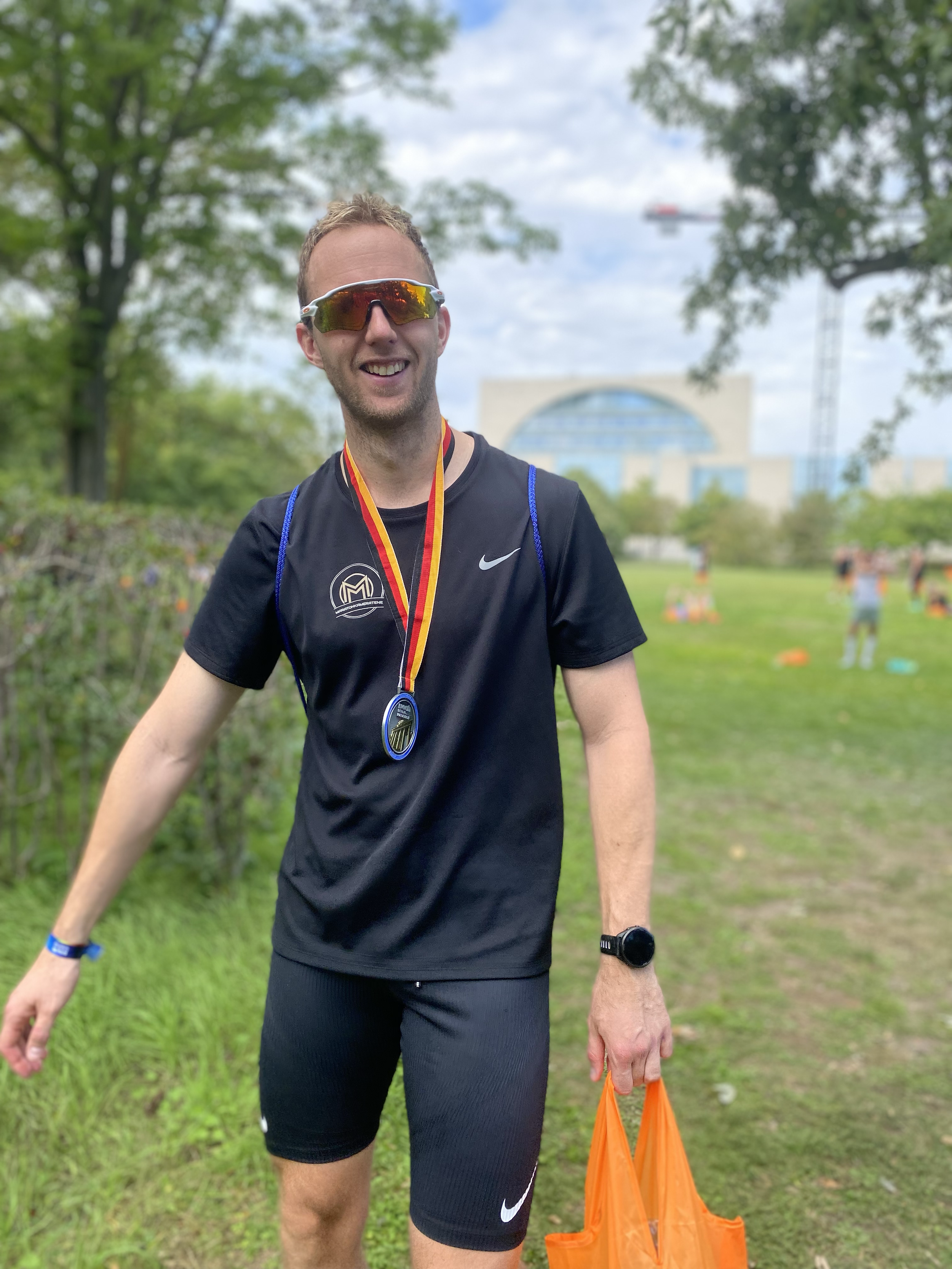 En løper med svart t-skjorte og shorts, med medalje rundt halsen, smilende, i en park med trær og en bygning i bakgrunnen.