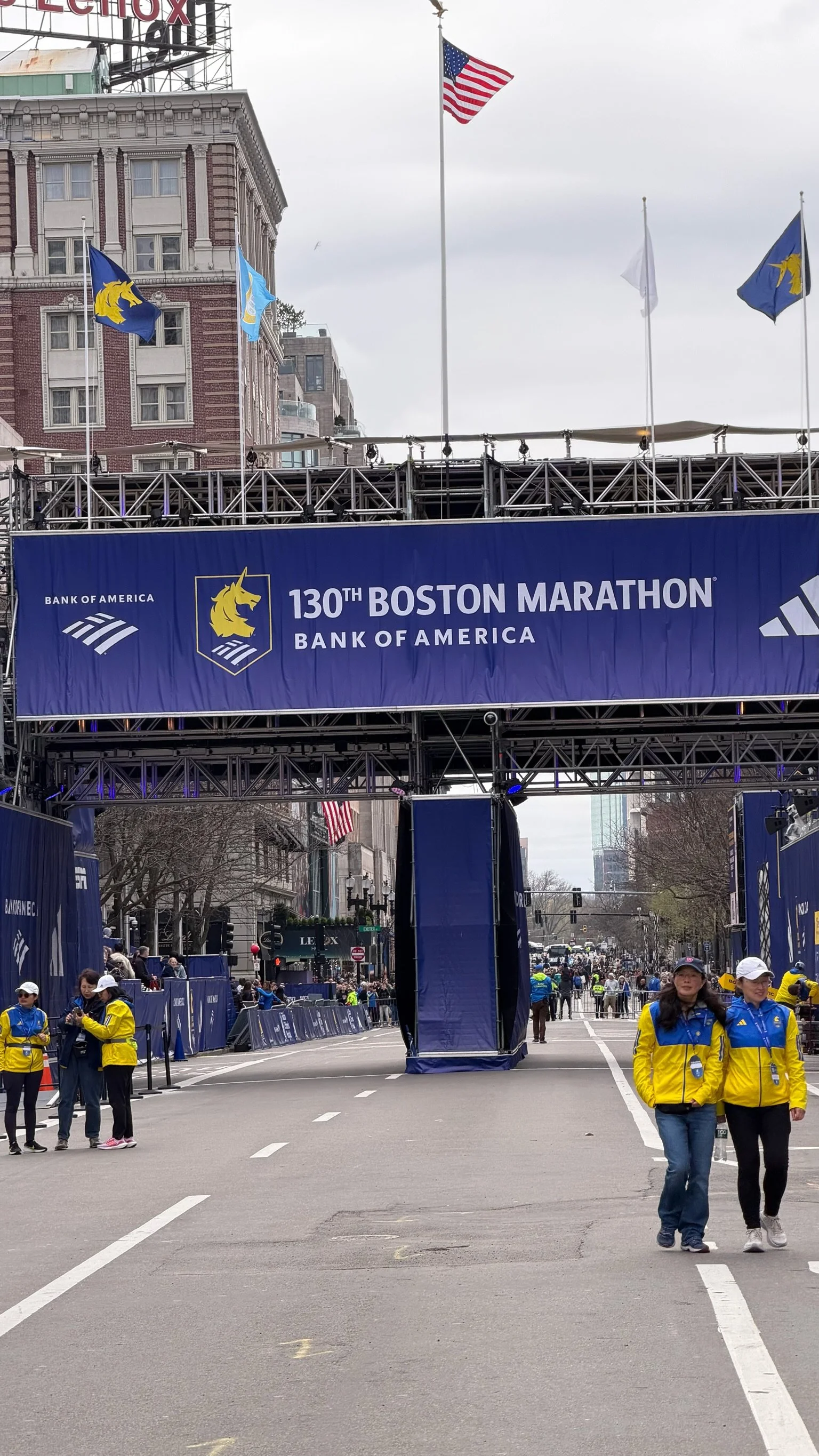 Start-området for 130. Boston Marathon med banner og arrangementspersonell iført gult og blått. Flere flagg, inkludert USAs flagg, vaier i bakgrunnen.