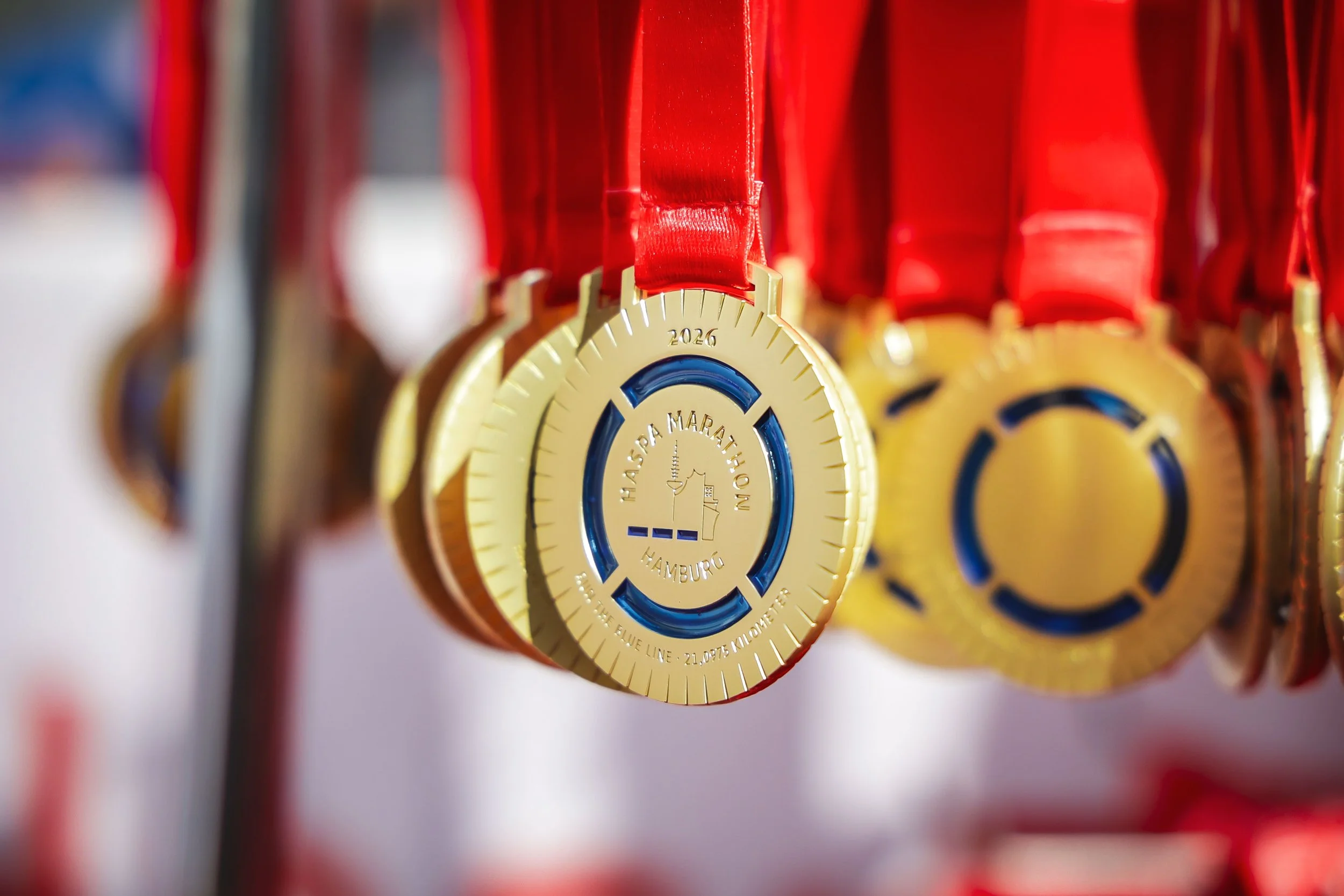 Nærbilde av gullmedaljer med røde bånd, hengende i en rekke. Det står «Hamburg Marathon 2026» på medaljene, og de har en blå sirkel i midten.