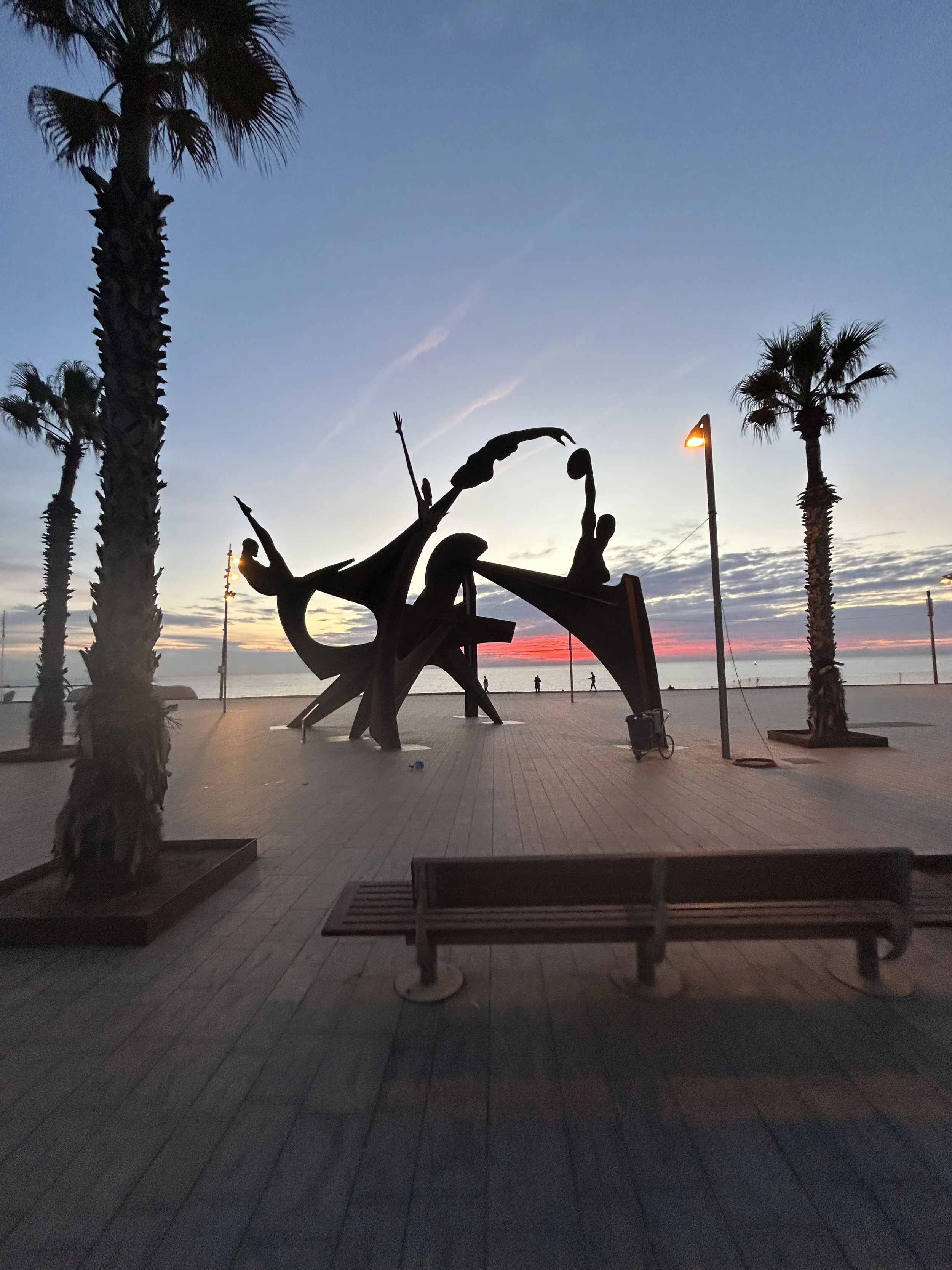 Siluett av skulpturer av mennesker som danser eller akker på en promenade ved havet, omgitt av palmetrær og lampelys, under en rosa og blå solnedgang.