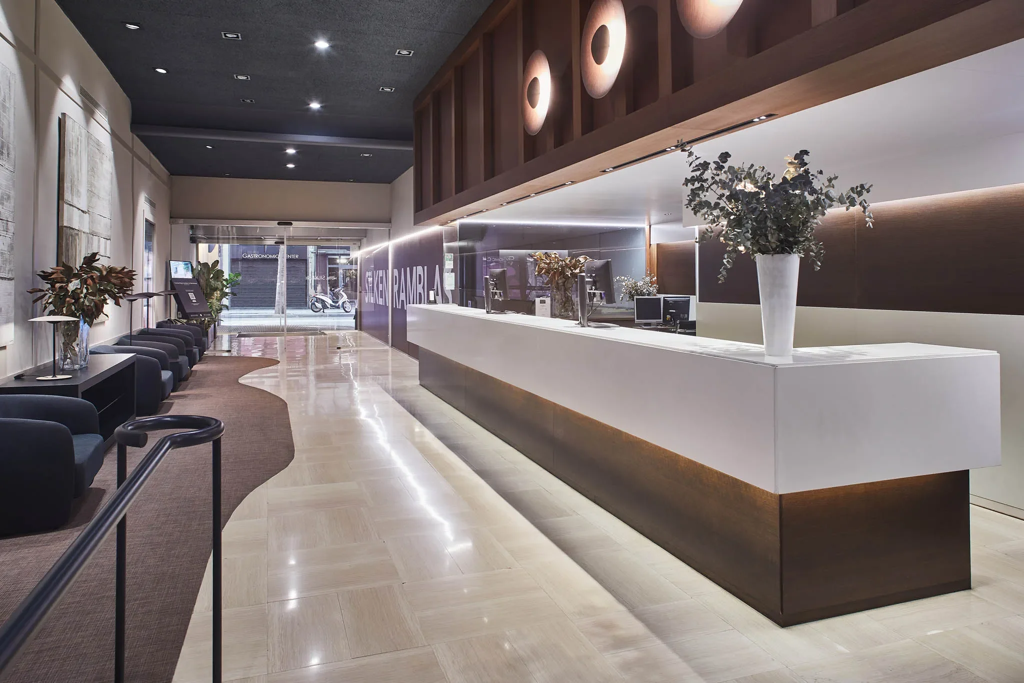 Resepsjonsområde med hvite og mørke møbler, grønne planter i store vaser, og en utsmykket mørk vegg. Avgangsknappene er synlige bak skranken, og det er store vinduer foran som viser inngangen.