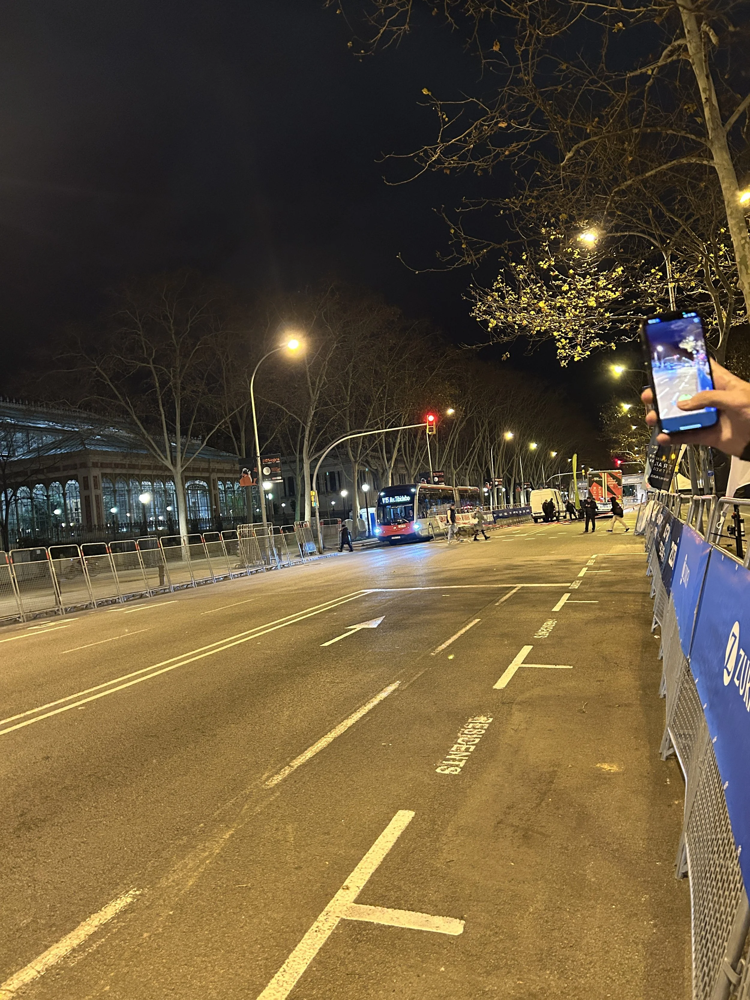 A street scene at night with a bus, traffic lights, and a person taking a photo with a smartphone. Barriers line the sidewalk, and trees without leaves are visible.