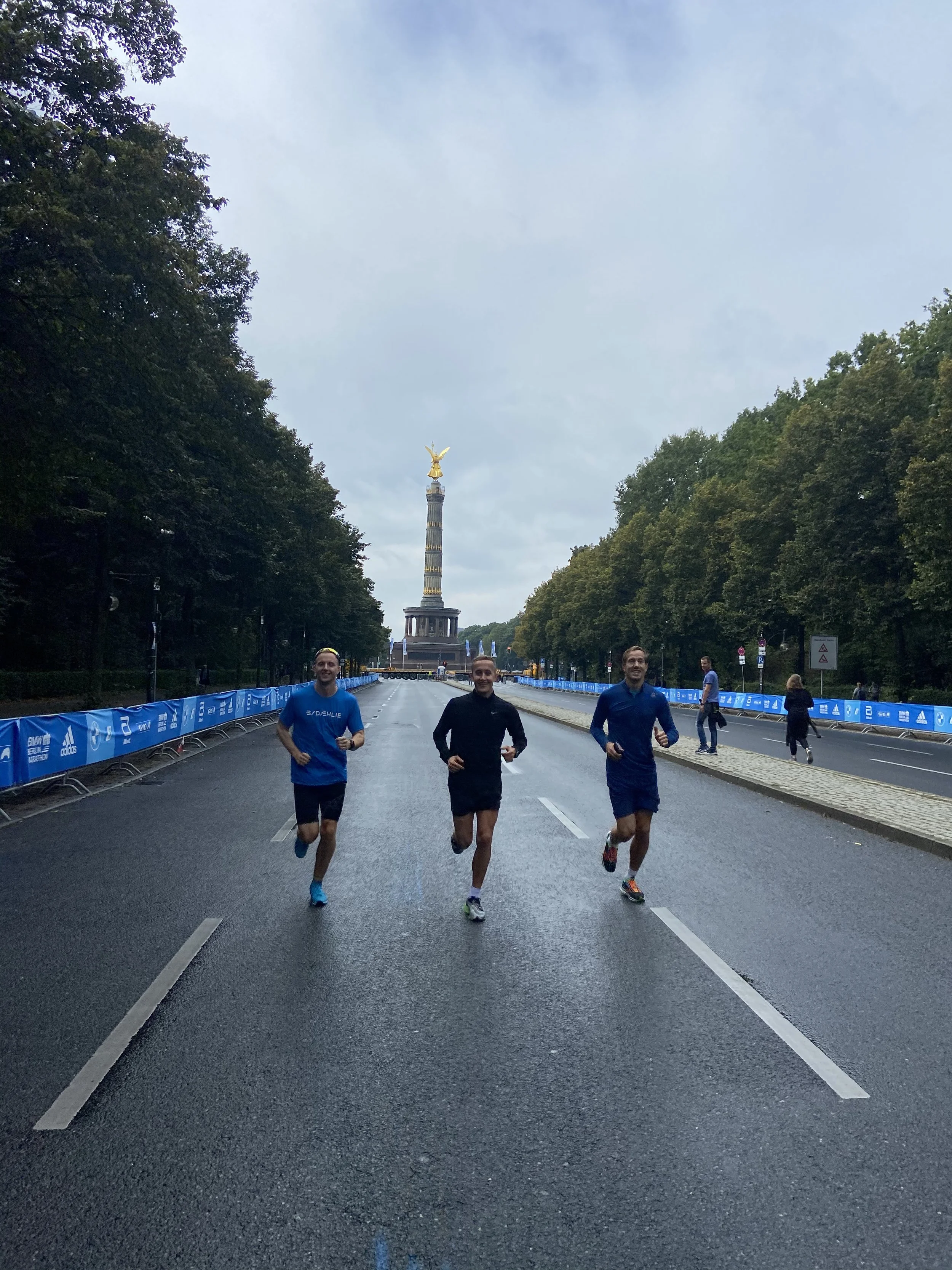 Tre løpere deltar i et løp, de løper på en nesten tom bygate med Berlinets milepæl, Siegessäule, i bakgrunnen. Det er overskyet, og veien er våt.