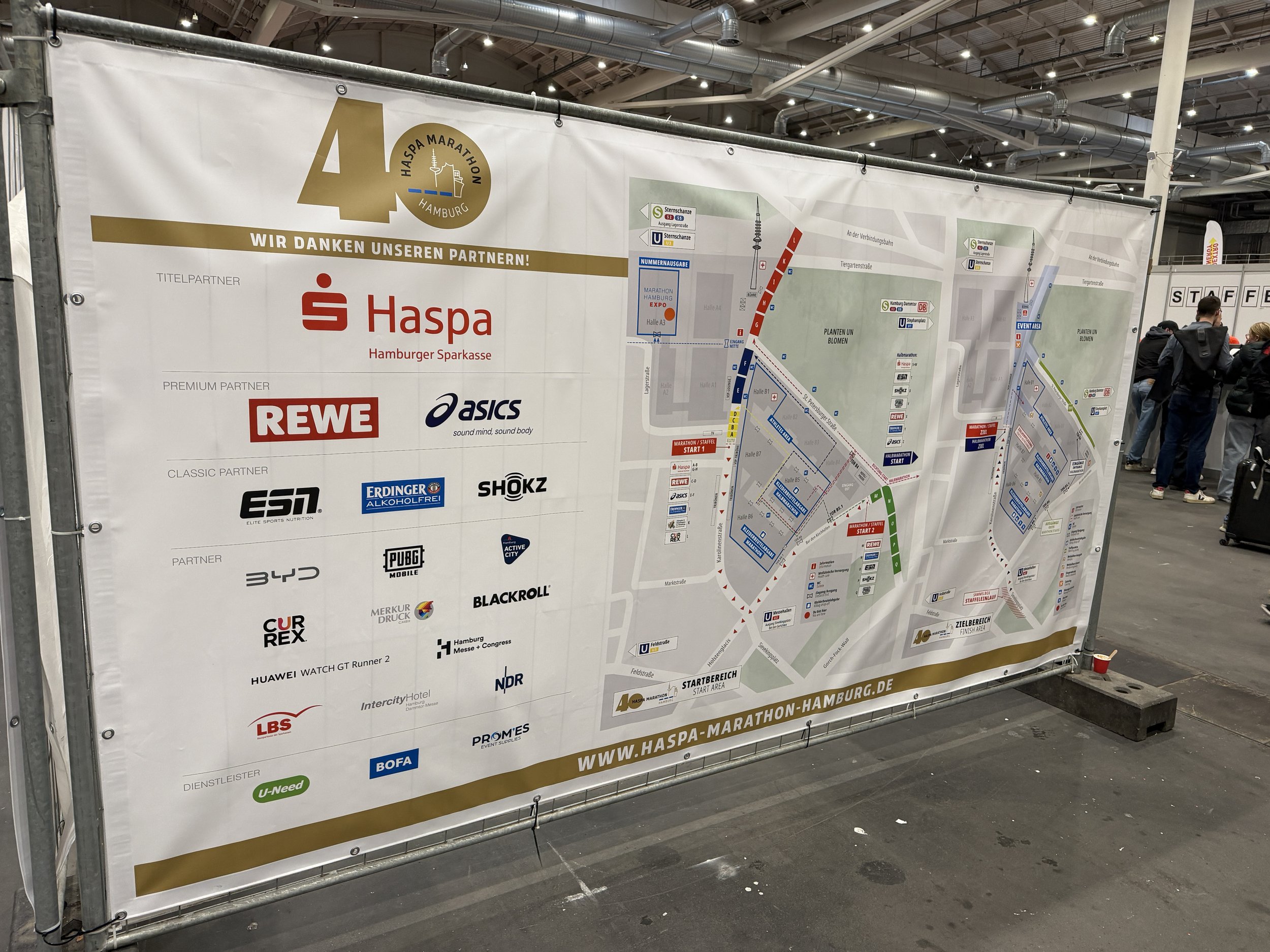 Informasjonsbanner ved et løpet som viser løypen og sponsorlogoer, inkludert Haspa Hamburg Marathon, med mennesker i bakgrunnen.