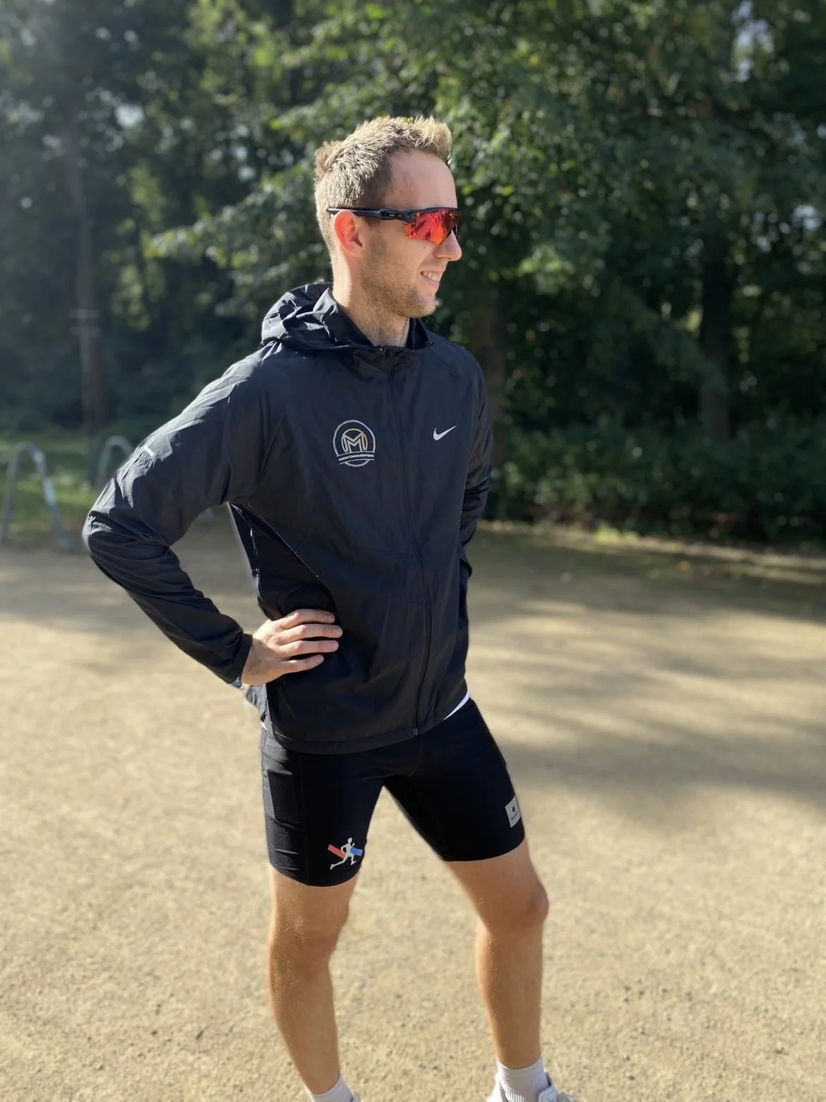 En mann i treningsklær står utendørs med hendene på hoftene, smilende og ser til høyre. Han bruker rød refleksbriller og en svart vindjakke med Nike-logo og noen andre logoer, samt svettebuksesko.