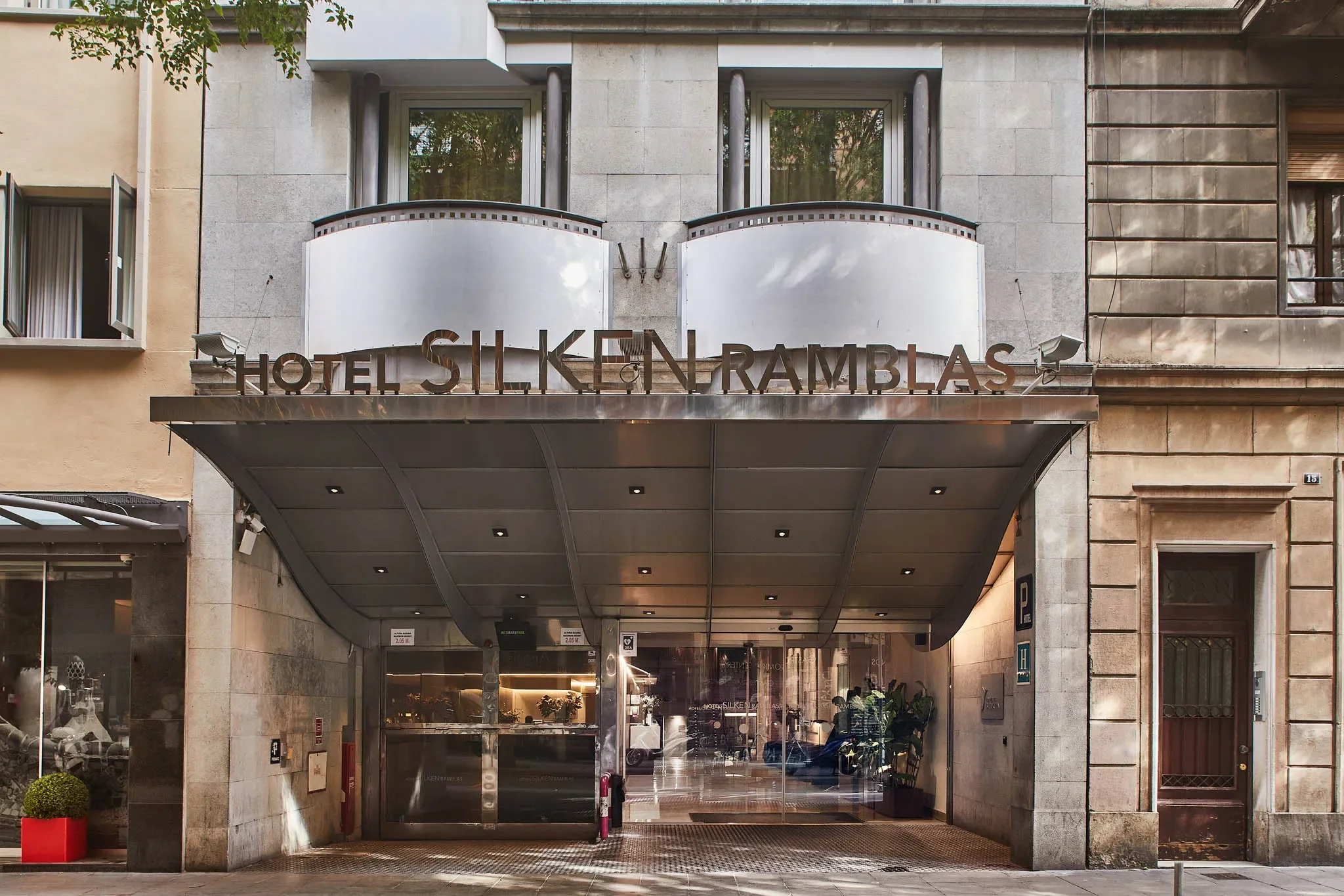 Inngangspartiet til hotellet Silken Rambla, med en inngangsdør under en overbygd takløkke, omgitt av bygninger i stein og glass, med skilt som viser hotellets navn.