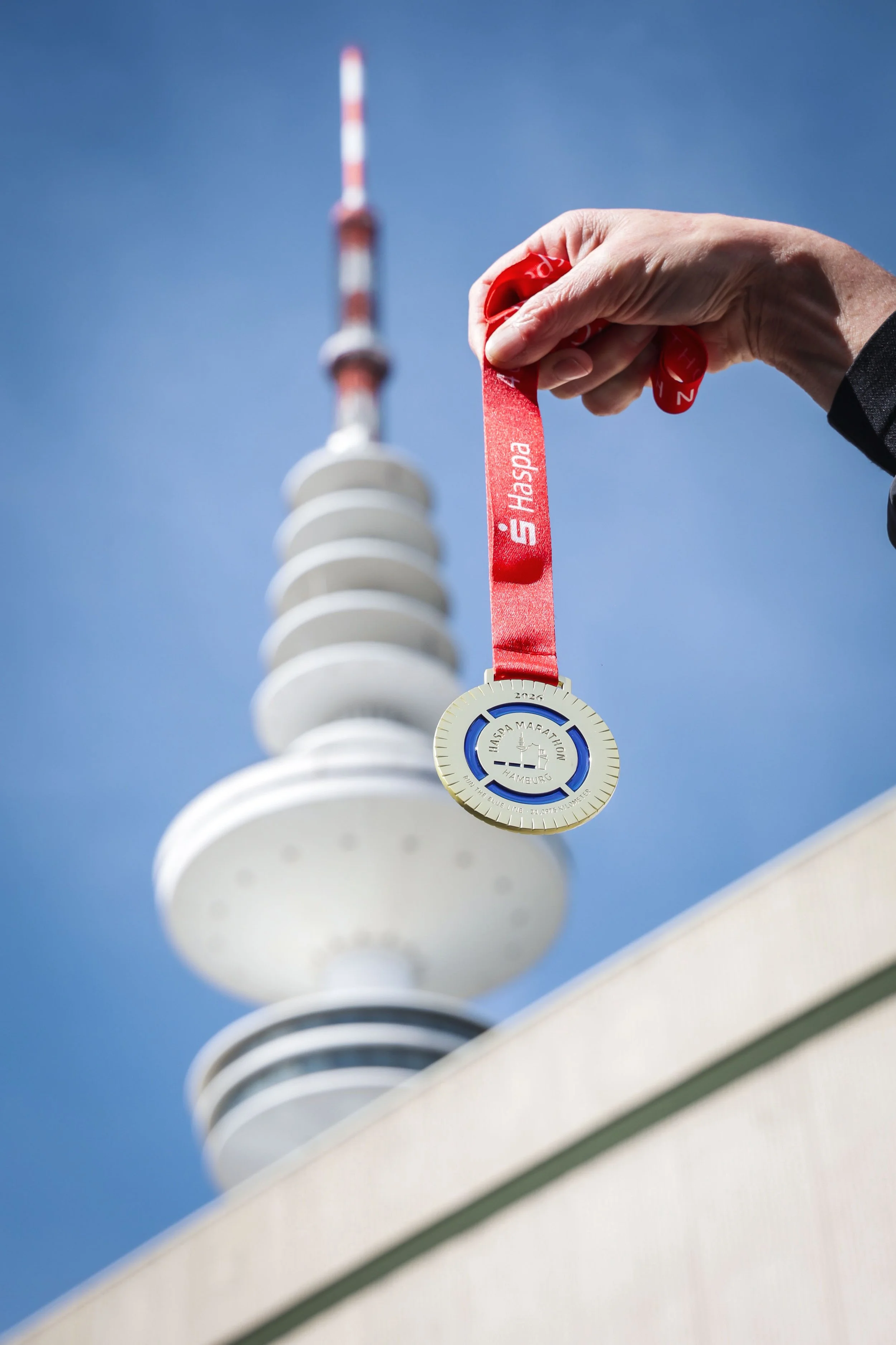 En person holder en gul medalje med tekst om et løp i Hamburg, med en bakgrunn av en klar blå himmel og en radiator.