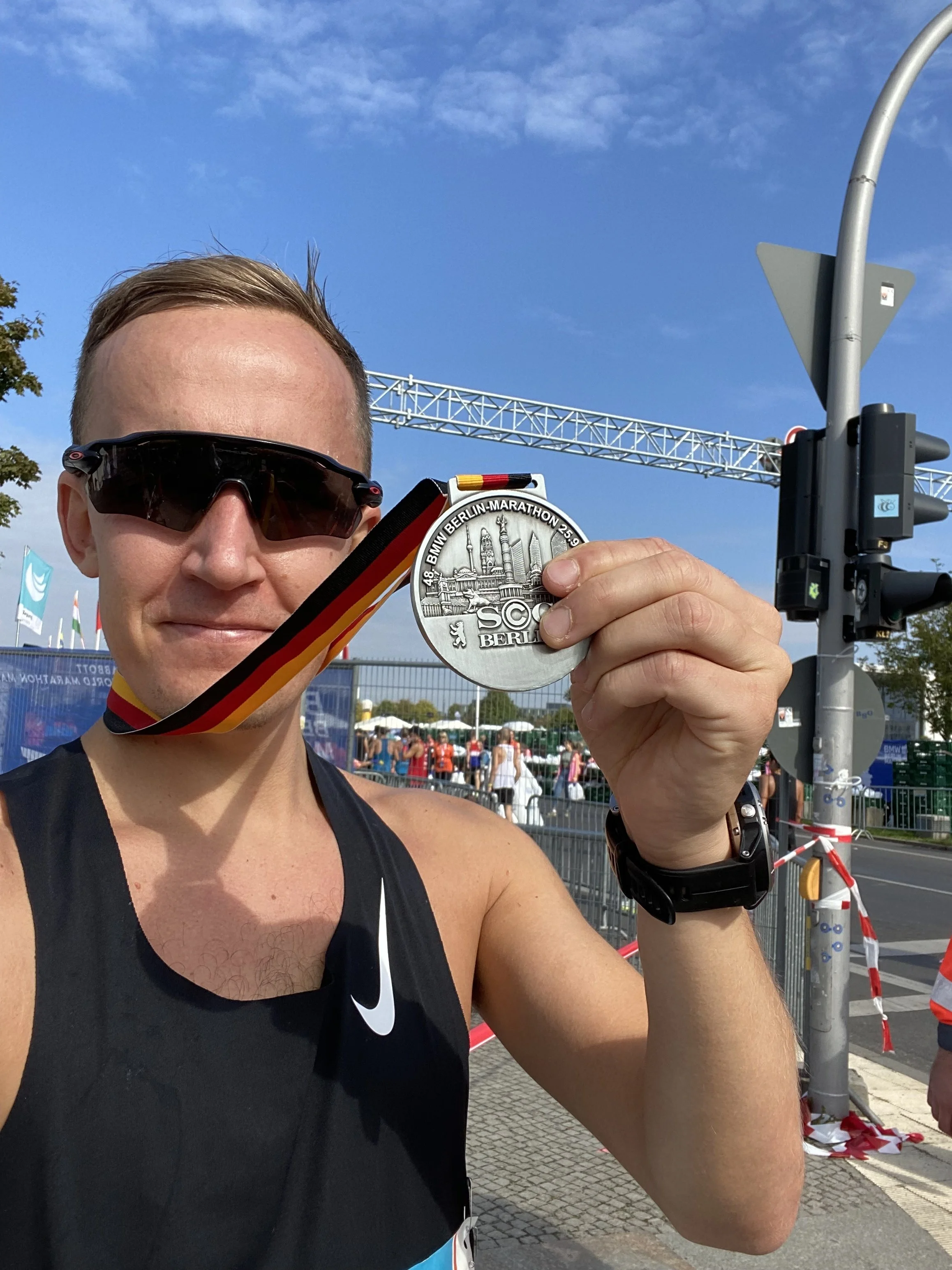 En mann med solbriller holder opp en sølvfarget medalje, som har teksten "BMW Berlin-Marathon 25.09.17" og et bilde av Berlin. Mannen er iført en svart Nike-treningsdrakt og står på en gate med trafikklys, og det er mange mennesker i bakgrunnen.