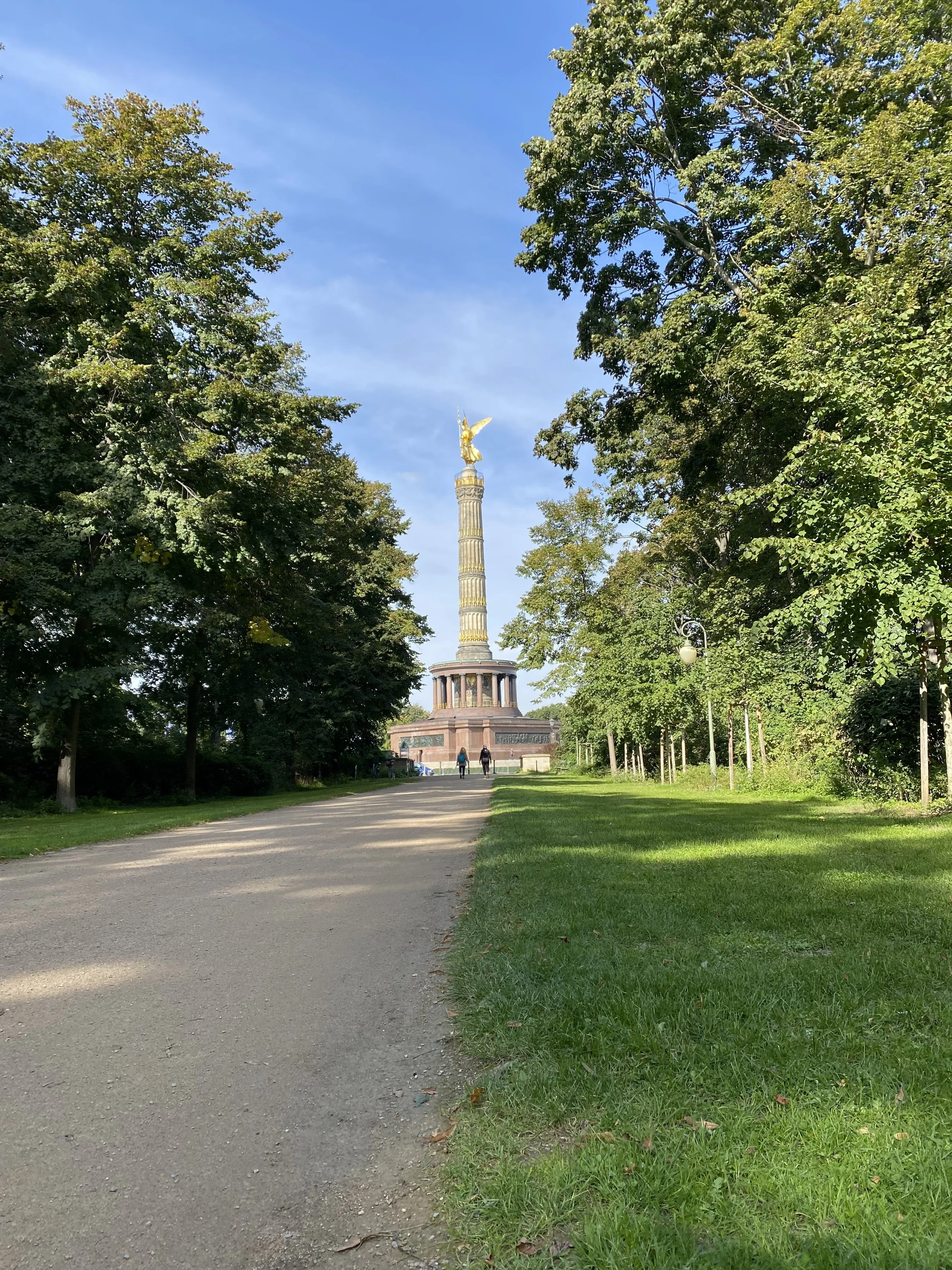 En park med en sti som går mot en kolonne med en gullfigur øverst, omgitt av grønne trær på klar dag.