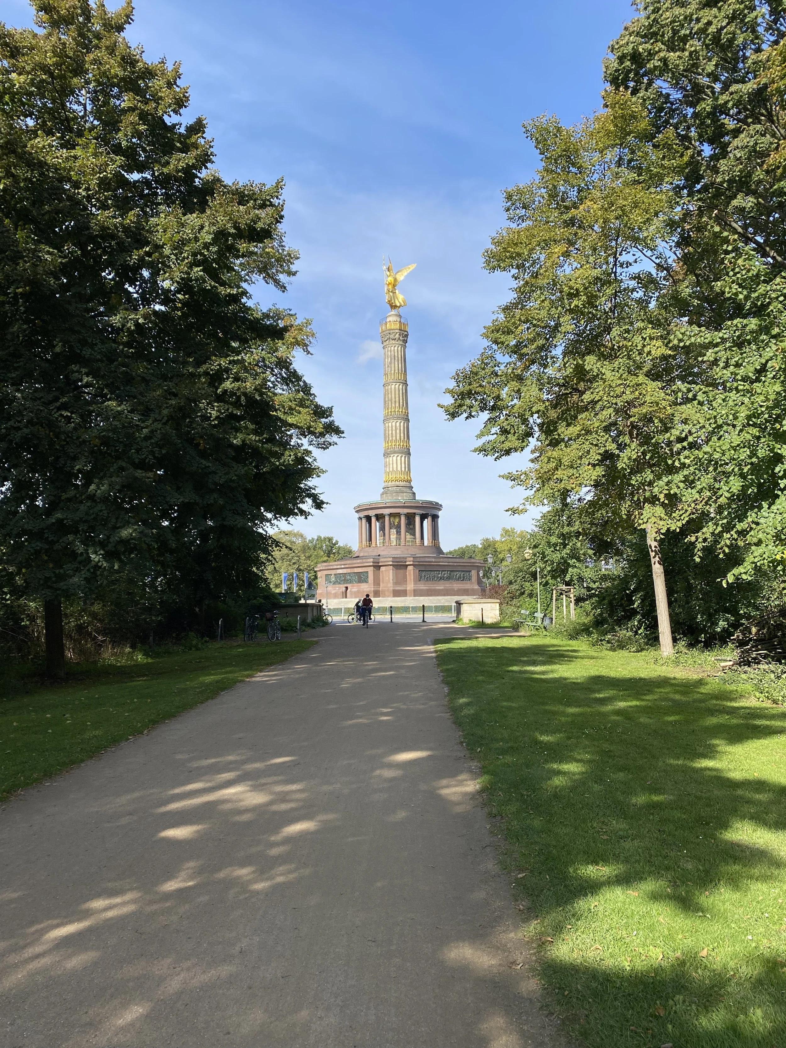 A park with a dirt pathway, lush green trees on both sides, and a tall monument with a golden angel at the top in the background.