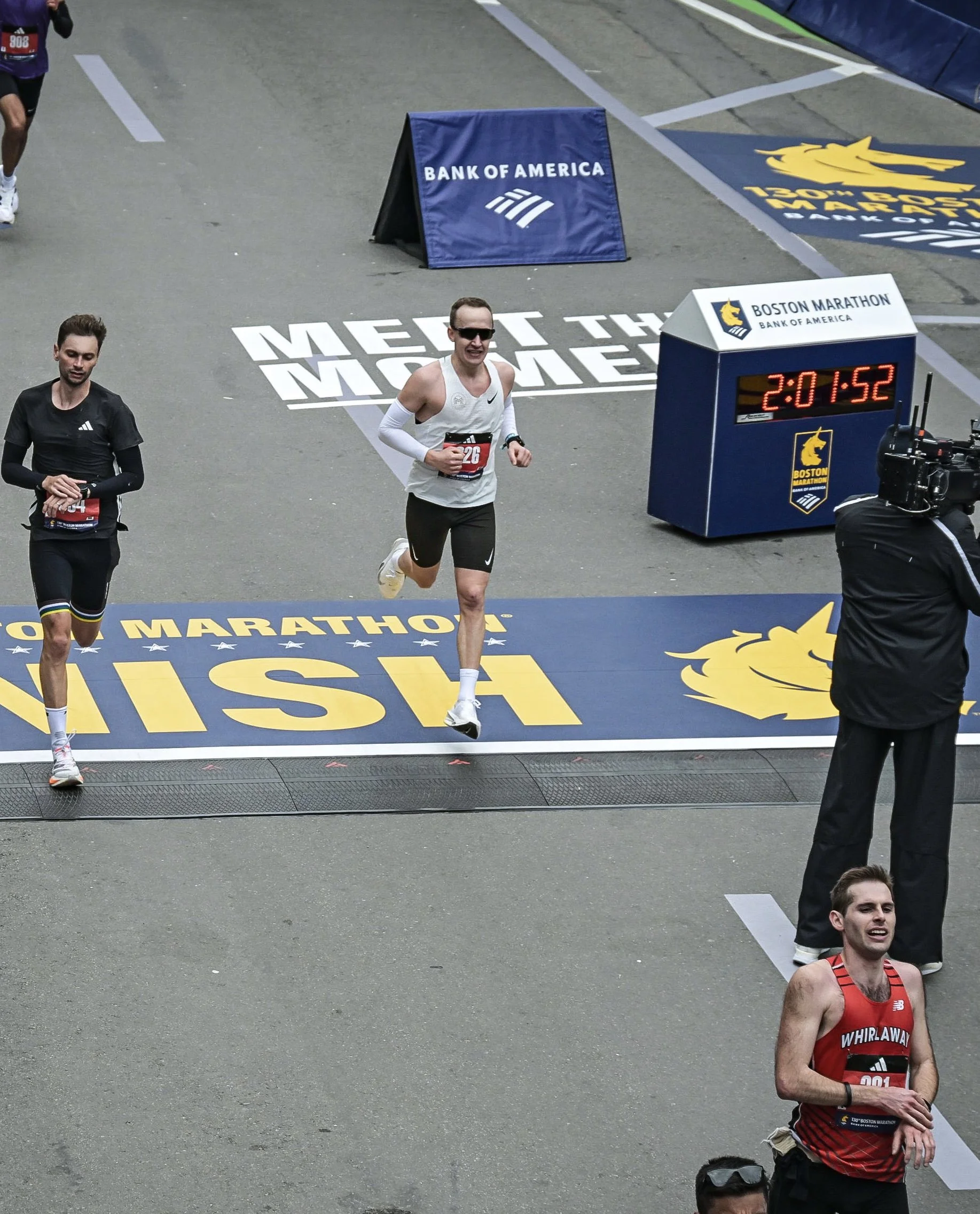 Maratonløpere som passerer målstreken ved Boston Marathon, med en digital klokke som viser tiden 2 timer, 1 minutt og 52 sekunder, omgitt av sponsormateriell som BANK OF AMERICA og Boston Marathon-logo.