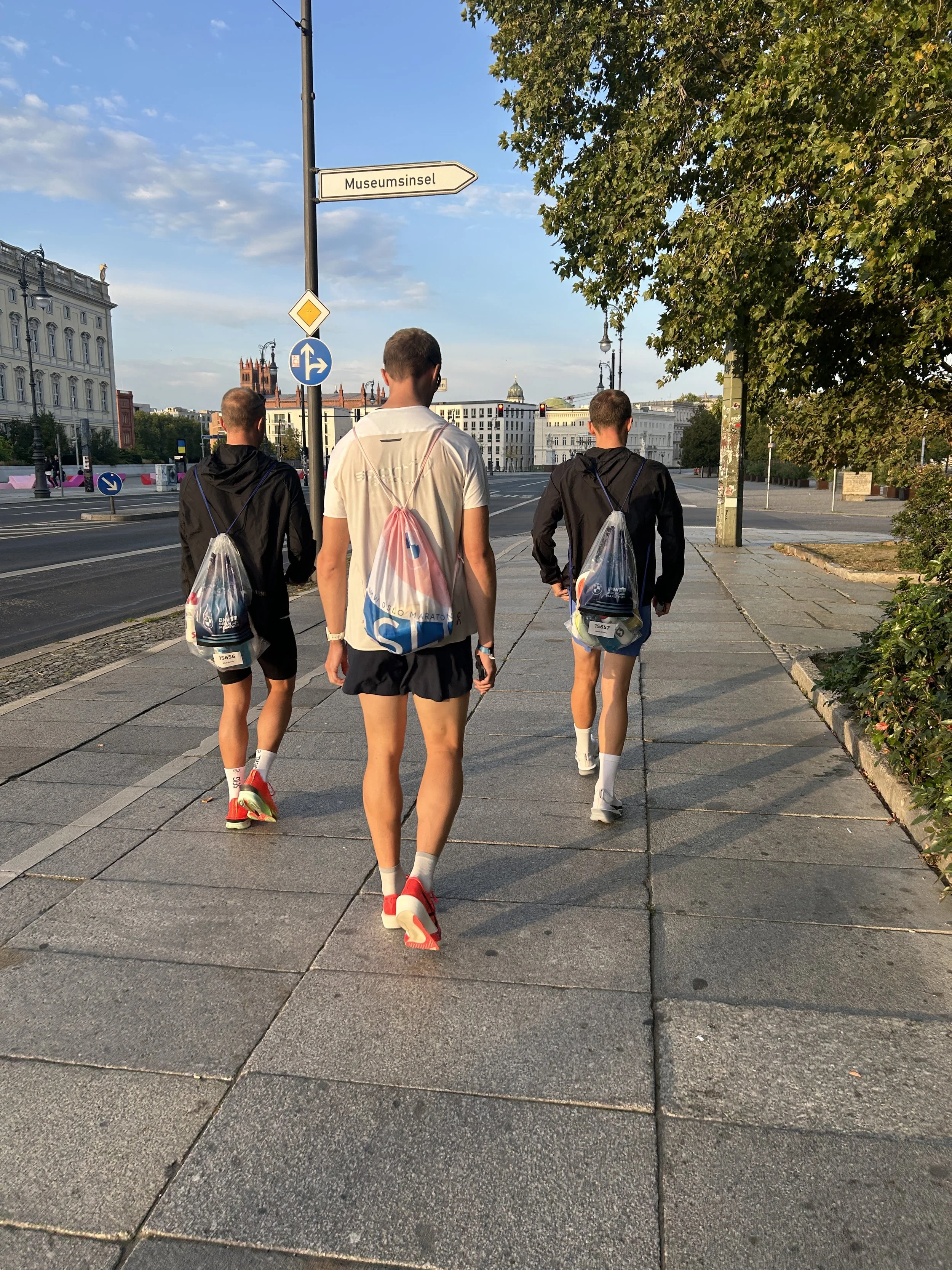 Tre personer går på fortauet, to menn i joggingklær og en i t-skjorte og shorts, alle med ryggsekker. Bak dem er det trafikksignaler og et bylandskap med gamle og moderne bygninger, en stor gravemaskin er også synlig.