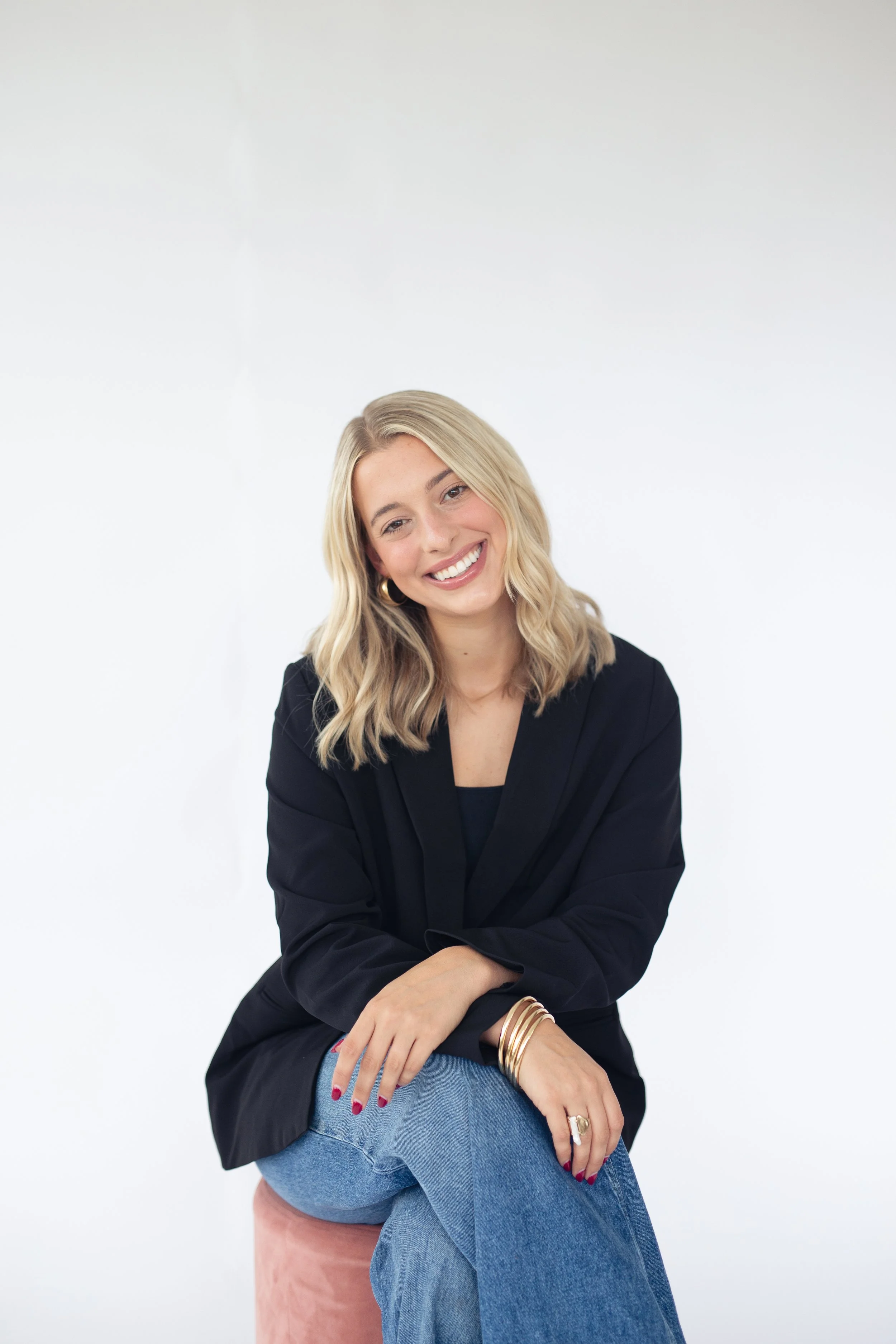 A young woman with blonde hair, smiling, sitting on a pink ottoman against a white background, wearing a black blazer, blue jeans, and gold jewelry.