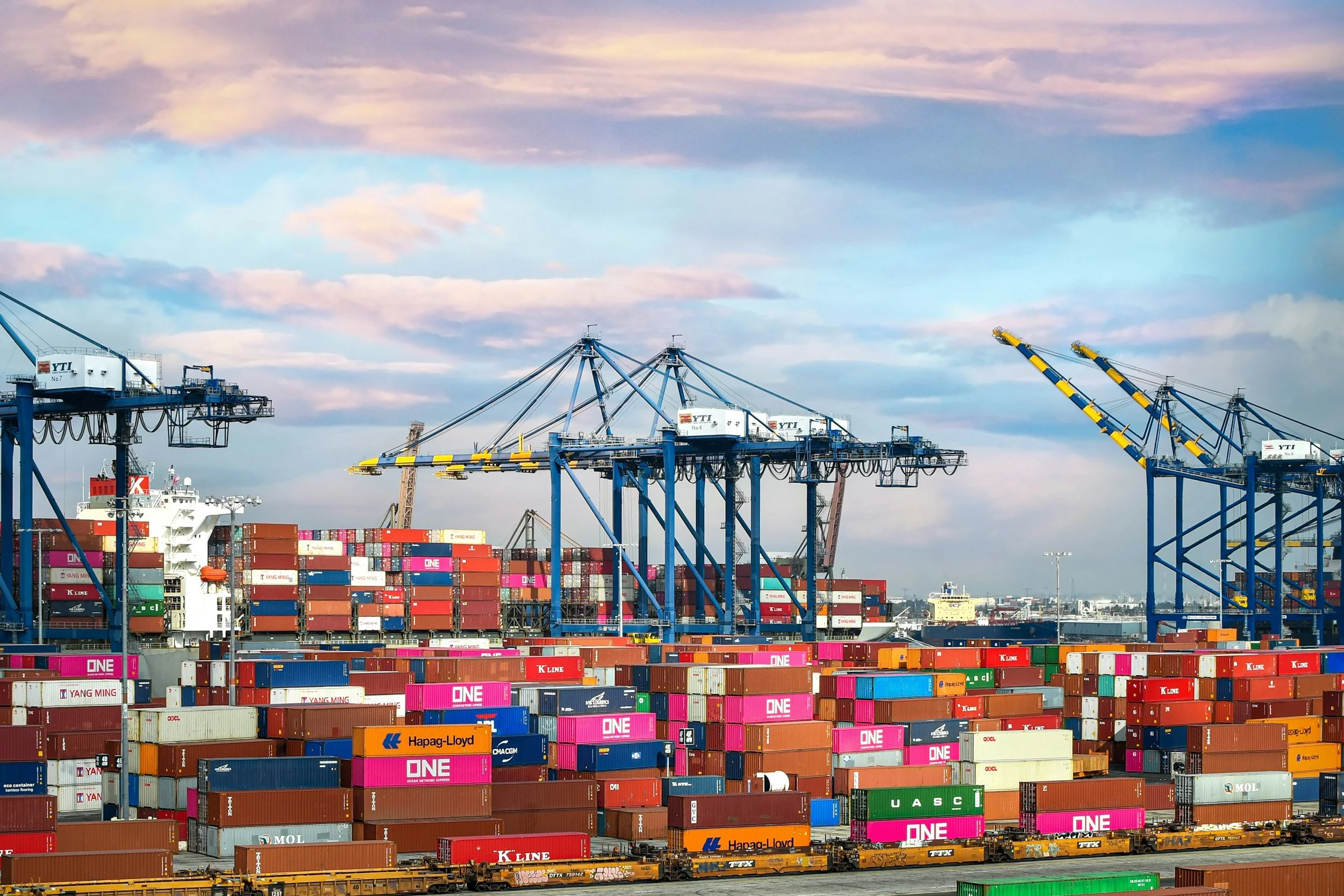 View of a busy container port with colorful shipping containers stacked, large blue cranes for loading and unloading, a partly cloudy sky, and railway trains in the foreground.