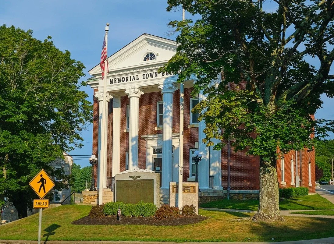 A historic brick building labeled 'Memorial Town Hall' with white columns, an American flag, and a small plaque in front. There are trees and a pedestrian crossing sign nearby.